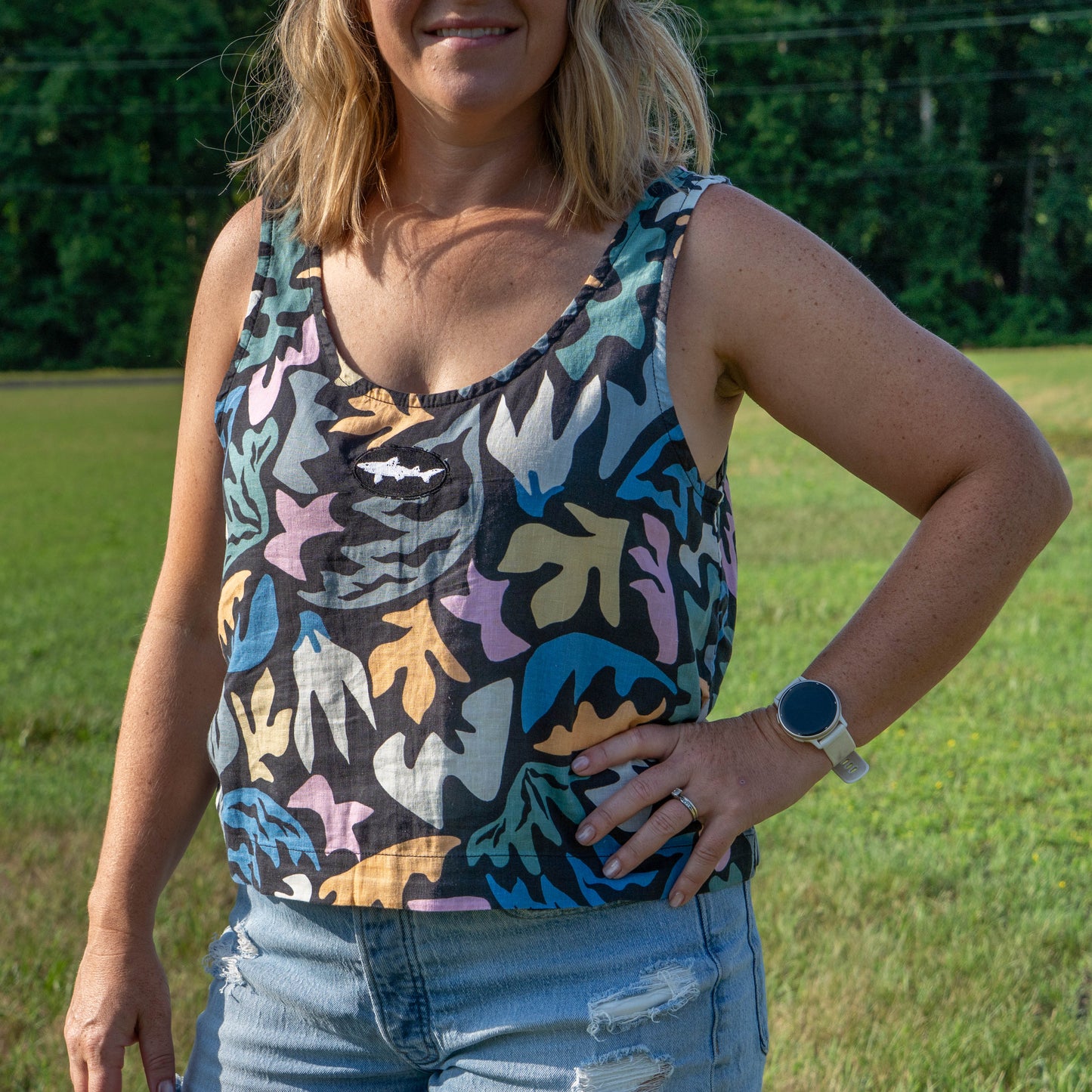 A woman stands on grass, wearing a Patagonia W Garden Island Tank Top with a botanical print, distressed denim shorts, and a smartwatch. Her hand is on her hip and there are trees in the background.