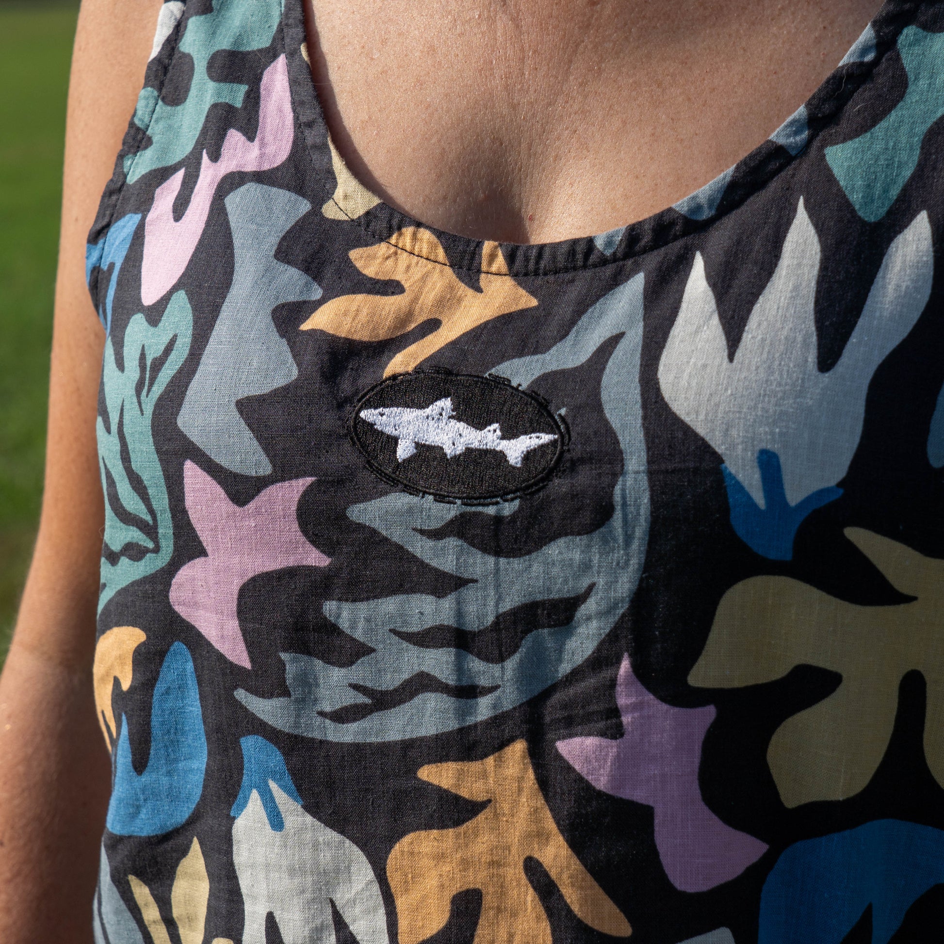 Wearing a vibrant Patagonia W Garden Island Tank Top, featuring a bold pattern and sleeveless design, someone stands out against a softly blurred grassy background, the white shark patch sewn on the chest drawing attention.