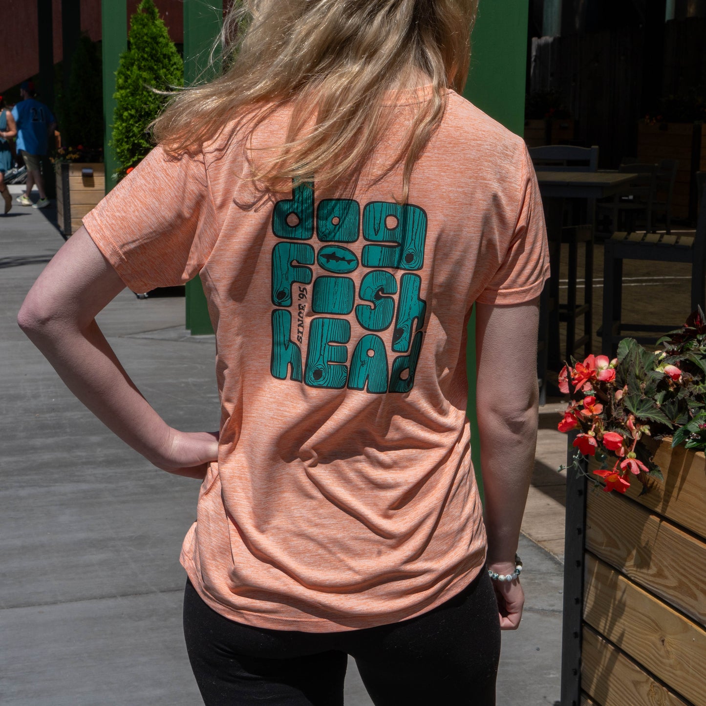 Back view of woman wearing Dogfish Head graphic on peach Patagonia Cap Cool Daily t-shirt, wood-textured blue-green lettering with dogfish shark emblem 