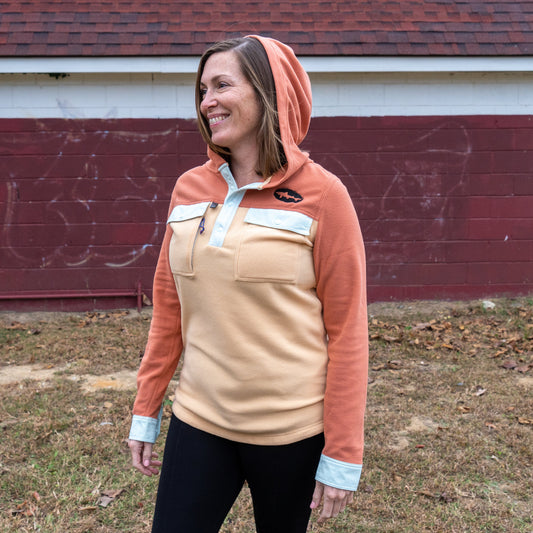 A woman wearing the Patagonia W Two Pocket Early Rise Pullover stands outdoors, smiling and looking to the side in front of a red and white painted building.