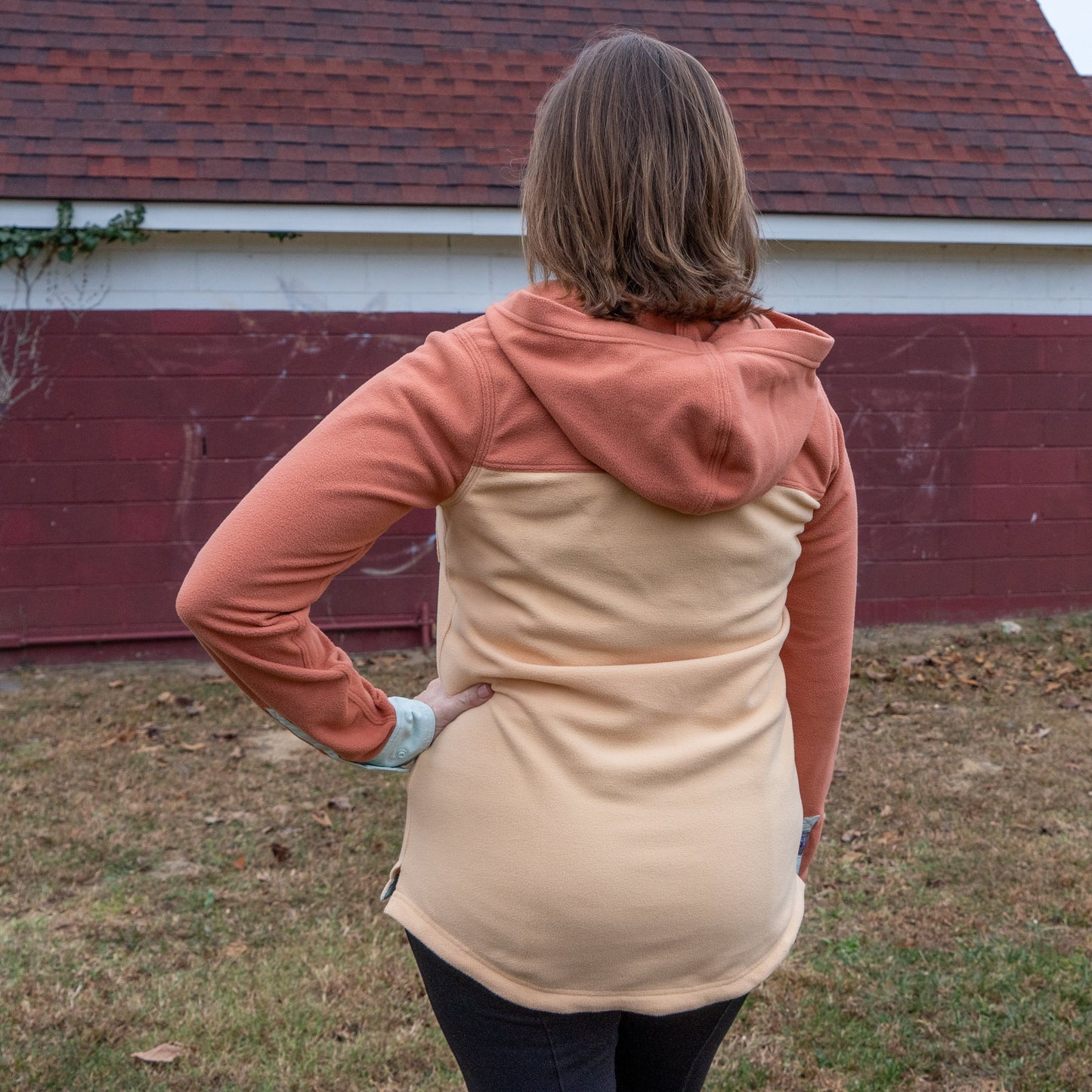 A person with light brown hair stands outside on grass, facing away, wearing the Patagonia W Two Pocket Early Rise Pullover by Patagonia. They are in front of a red and white building with a shingled roof.