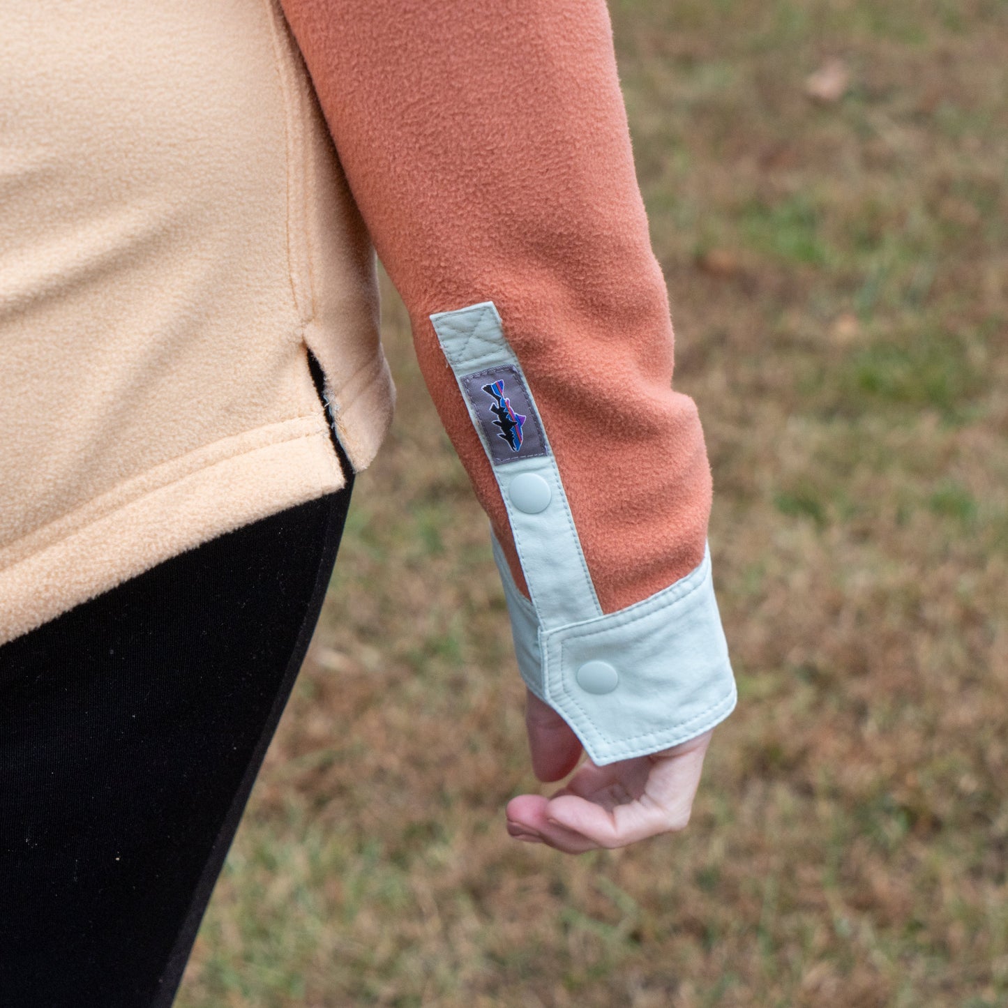 A person wearing the Patagonia W Two Pocket Early Rise Pullover, a peach and beige fleece jacket with light blue cuffs and snap buttons, stands outdoors on grass with only their arm and part of their torso visible.