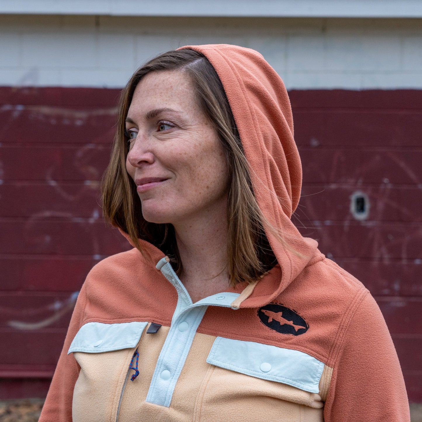 A woman with light brown hair stands outdoors by a red brick wall with white graffiti, smiling slightly to the side as she wears the Patagonia W Two Pocket Early Rise Pullover in orange, featuring a fish logo.