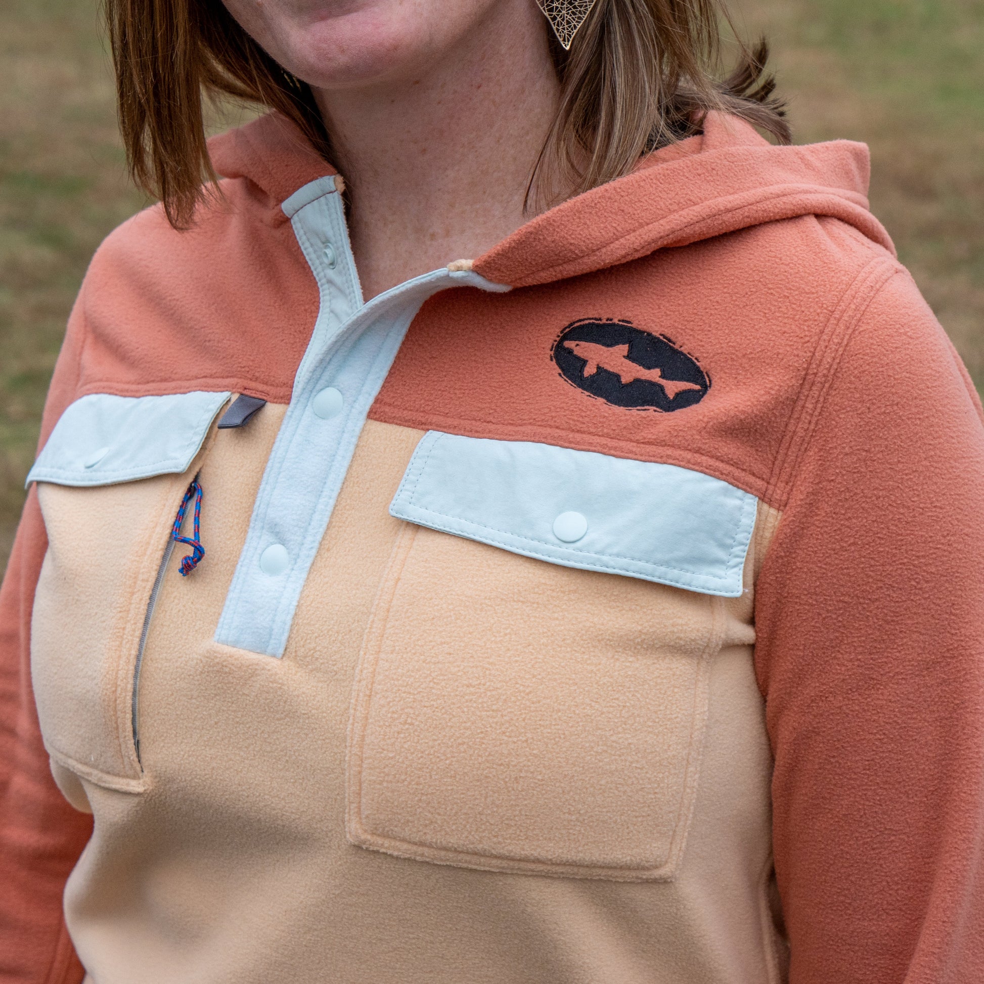 A woman wears the Patagonia W Two Pocket Early Rise Pullover by Patagonia, featuring peach and tan two-tone fleece, light blue chest pockets, and a fish-shaped shoulder patch. The image shows her upper torso and part of her face.