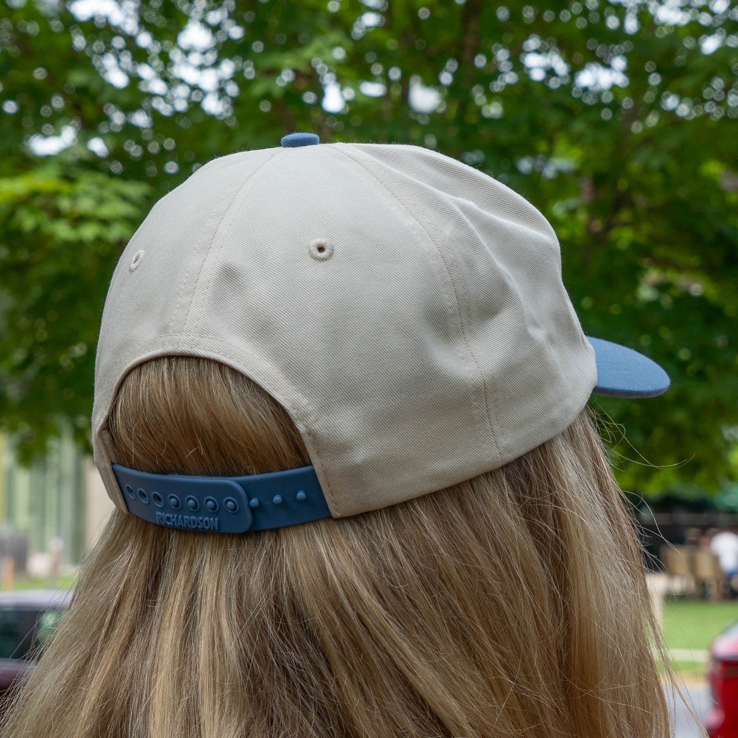 Person wearing a beige cap with blue brim and back, standing outdoors with greenery in the background.