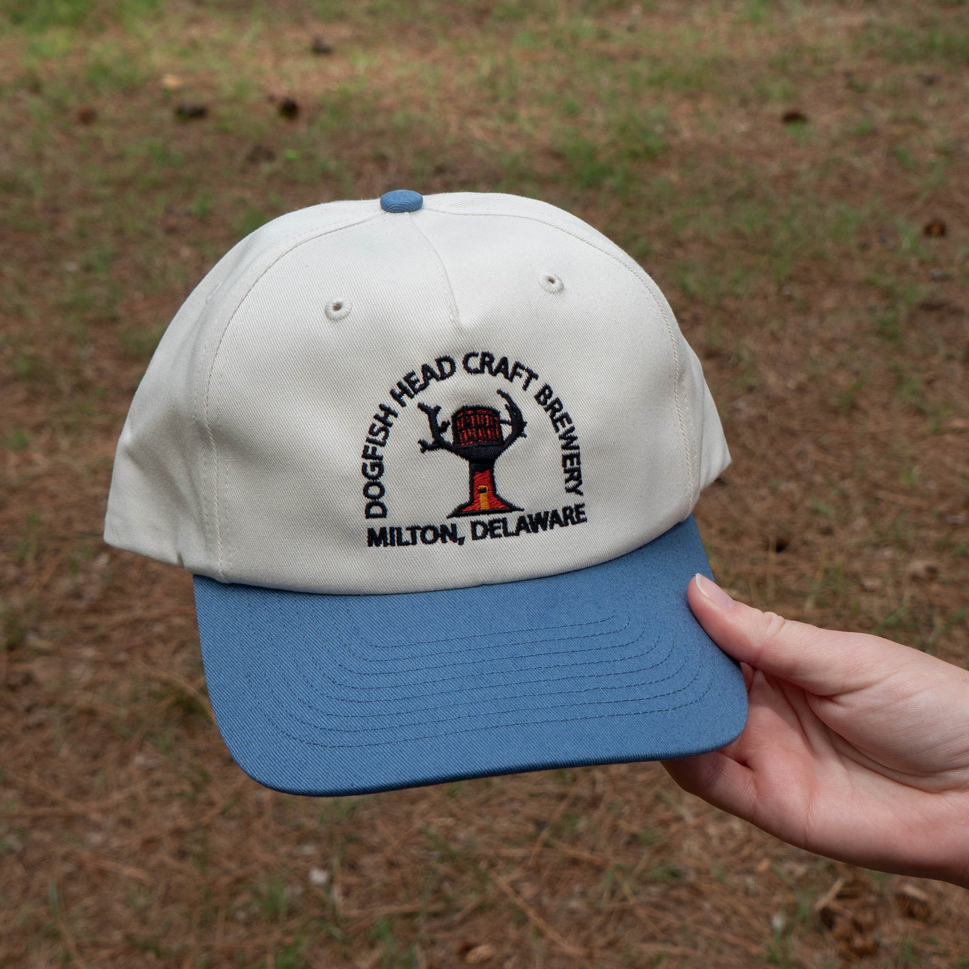 White cap with blue brim featuring Dogfish Head Craft Brewery logo held by a hand against a grassy background.