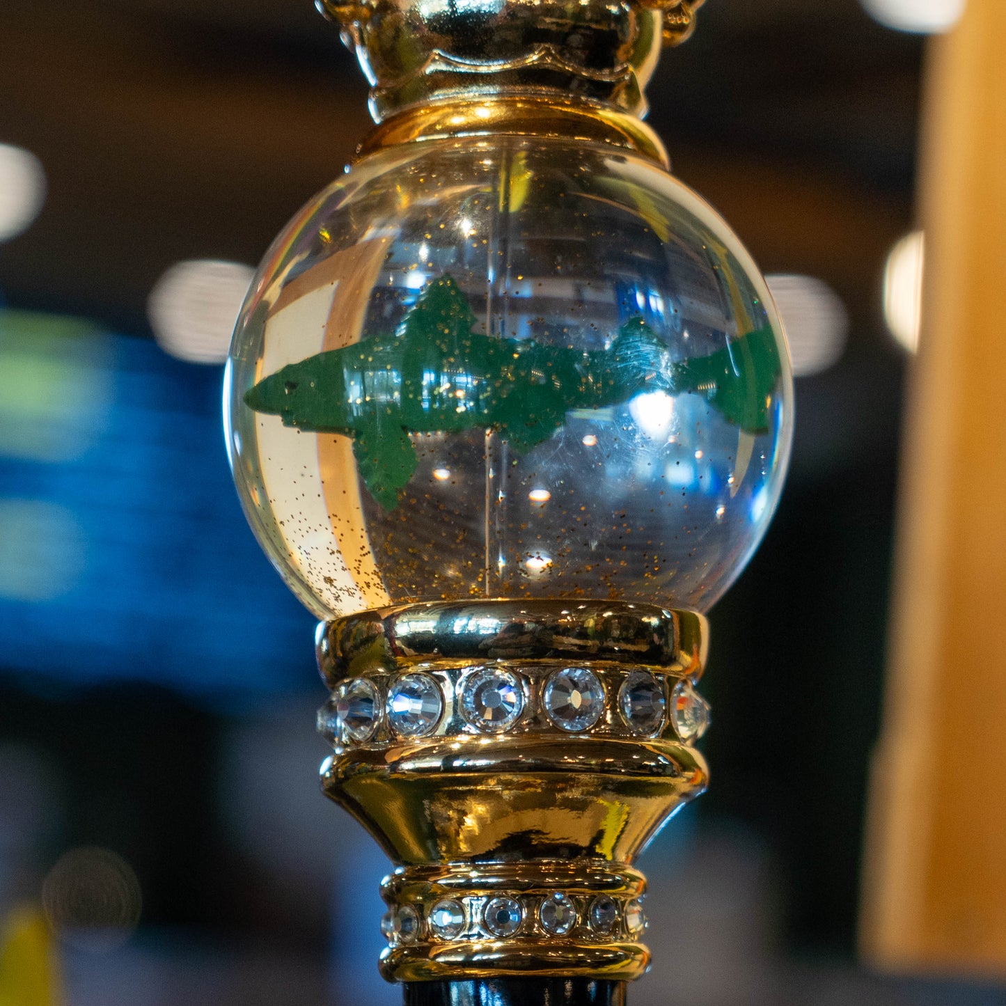 The OG Uber Tap Handle by Taphandles Inc. features a clear glass orb with a green shark inside, set on a gold jeweled stand, making it a true collectible tap handle for enthusiasts. The softly blurred background adds to its appeal.