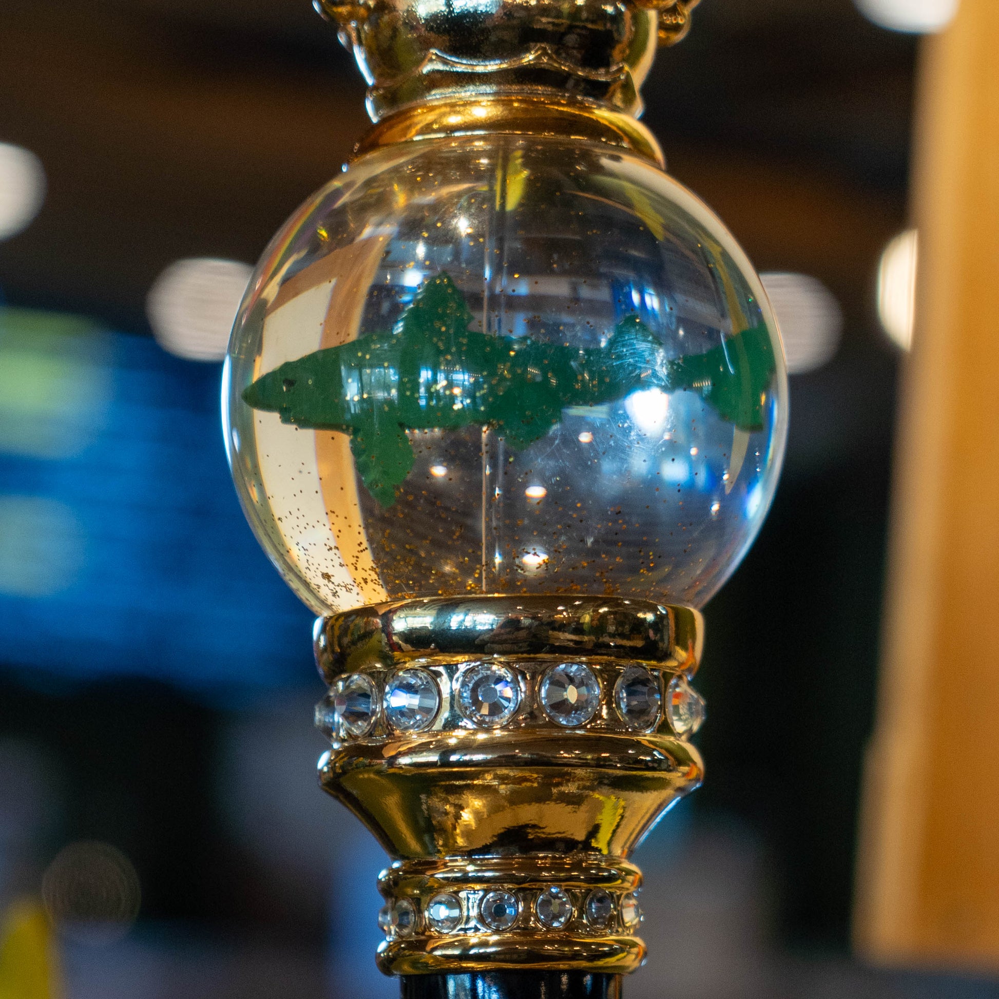 The OG Uber Tap Handle by Taphandles Inc. features a clear glass orb with a green shark inside, set on a gold jeweled stand, making it a true collectible tap handle for enthusiasts. The softly blurred background adds to its appeal.