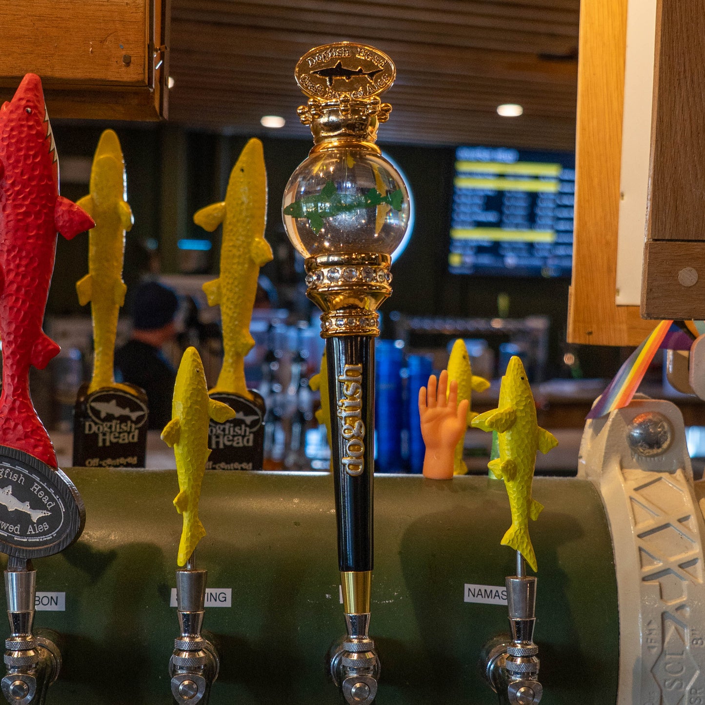 A row of beer taps at a bar features three collectible Dogfish Head tap handles with yellow fish designs and a central OG Uber Tap Handle by Taphandles Inc., crafted from ornate gold and glass, with a menu board and the bar's interior in the background.
