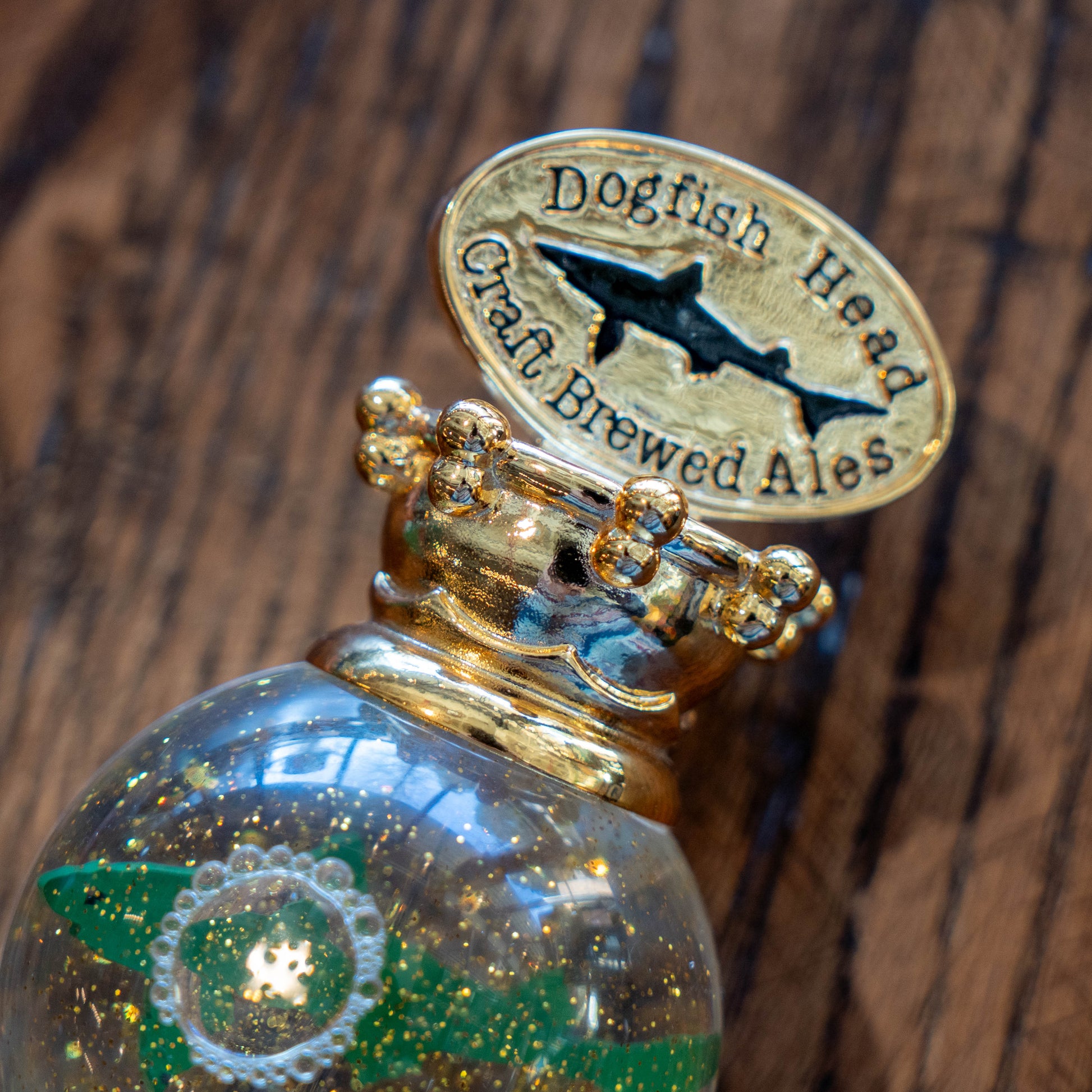 Close-up of the OG Uber Tap Handle by Taphandles Inc.—a gold, ornate stopper embossed with Dogfish Head Craft Brewed Ales and a shark logo, featuring gold flecks, attached to a bottle on a wooden surface.
