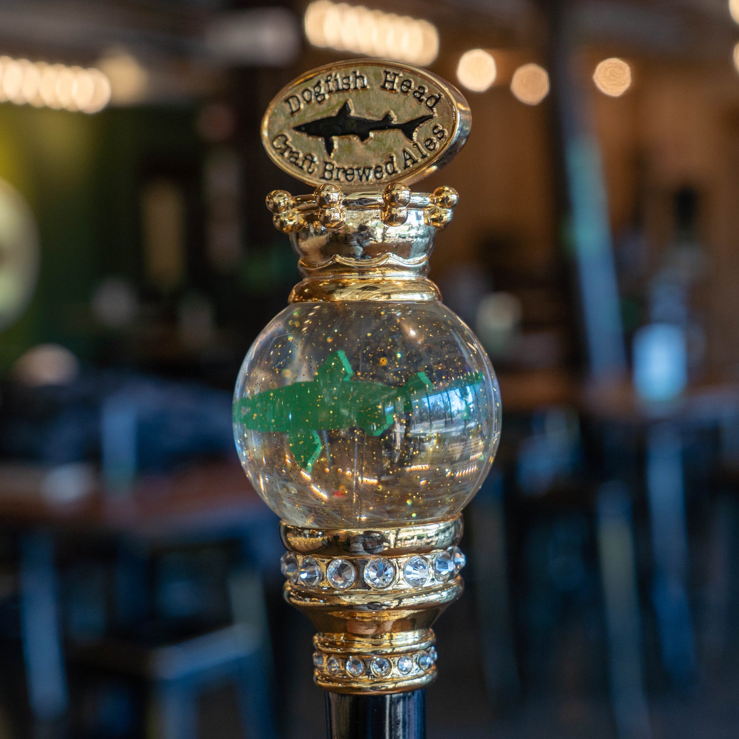 A close-up of the OG Uber Tap Handle by Taphandles Inc., featuring a clear globe with a green shark and sparkles, labeled Dogfish Head Craft Brewed Ales, set against a blurred bar background.