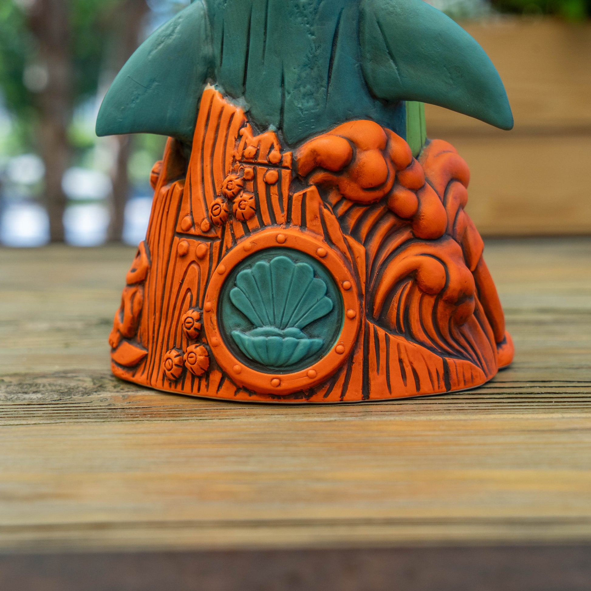 A close-up of a shark shaped ceramic cup with a teal top and an orange sculpted base featuring wood-grain texture, raised shell motifs, and wave-like patterns, placed on a wooden surface with a blurred outdoor background.