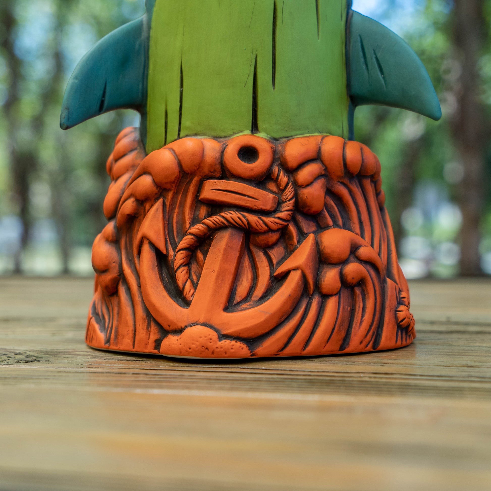 A close-up of a ceramic shark ceramic tiki mug base featuring a carved orange anchor and nautical wave pattern, with a green and teal tiki-style handle visible behind it, set on a wooden surface with a blurred outdoor background.