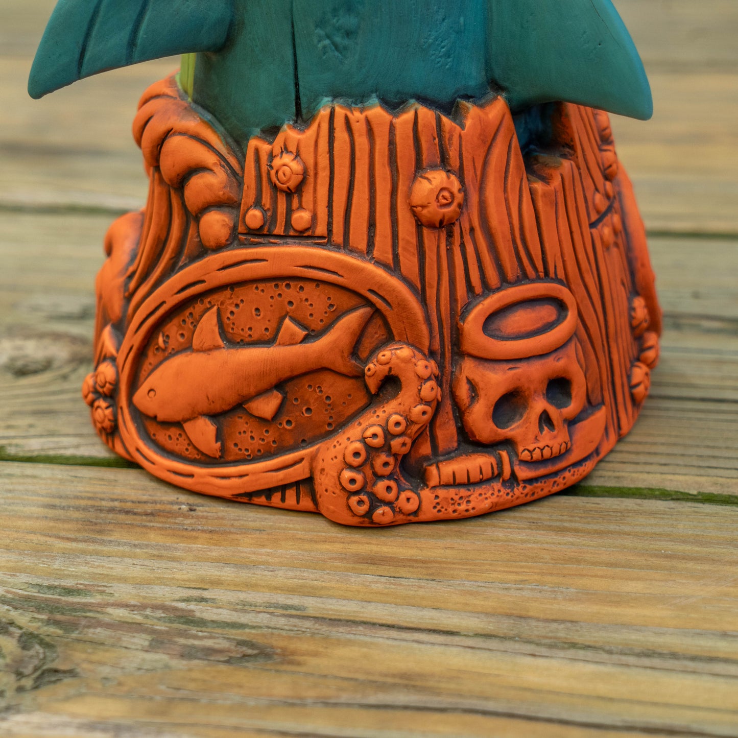 An orange carved base of a ceramic shark cup with a fish, skull, and tentacle motif, shown on a weathered wooden surface with part of a teal object visible at the top.