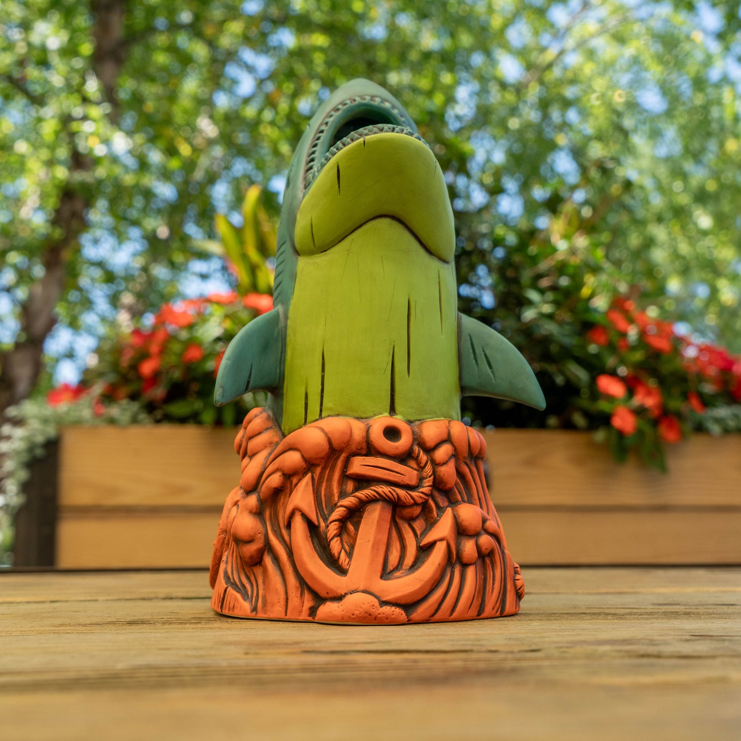 A ceramic shark cup with a green body and dark teal fins, standing on an orange nautical base with an anchor and rope, photographed outdoors on a wooden surface with blurred greenery and flowers in the background.