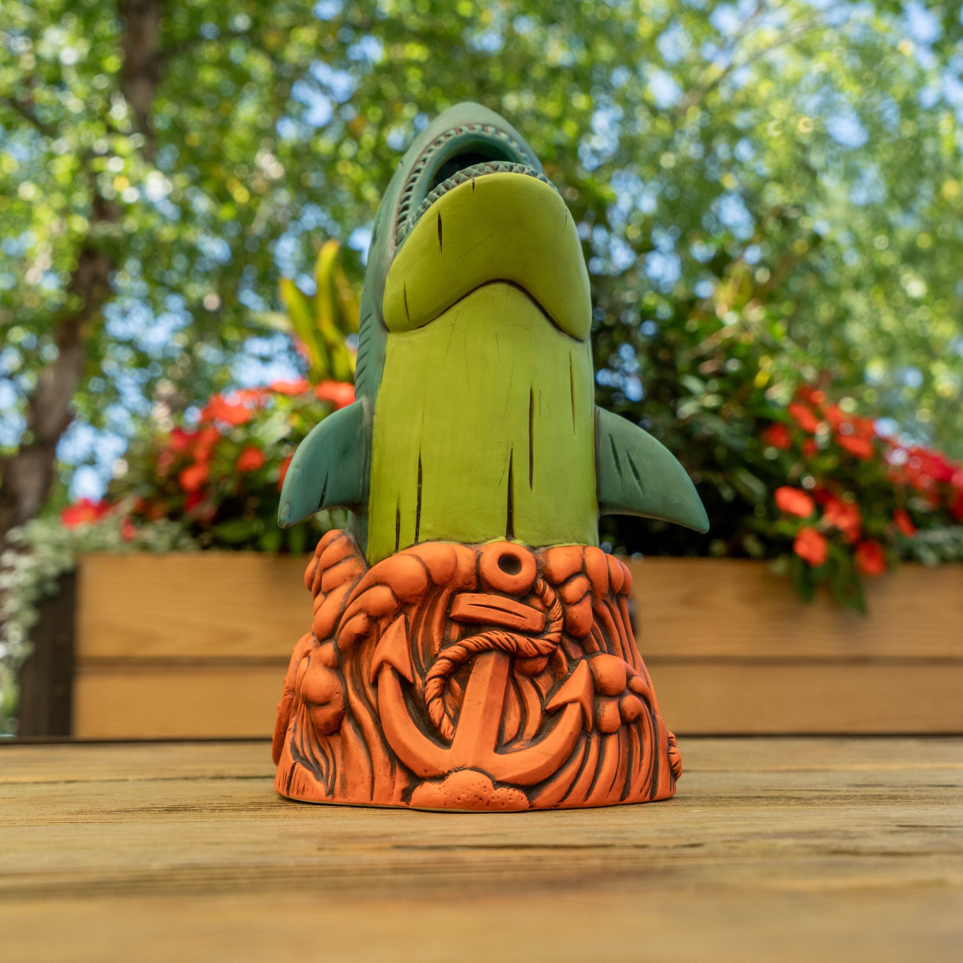 A ceramic shark cup with a green body and dark teal fins, standing on an orange nautical base with an anchor and rope, photographed outdoors on a wooden surface with blurred greenery and flowers in the background.