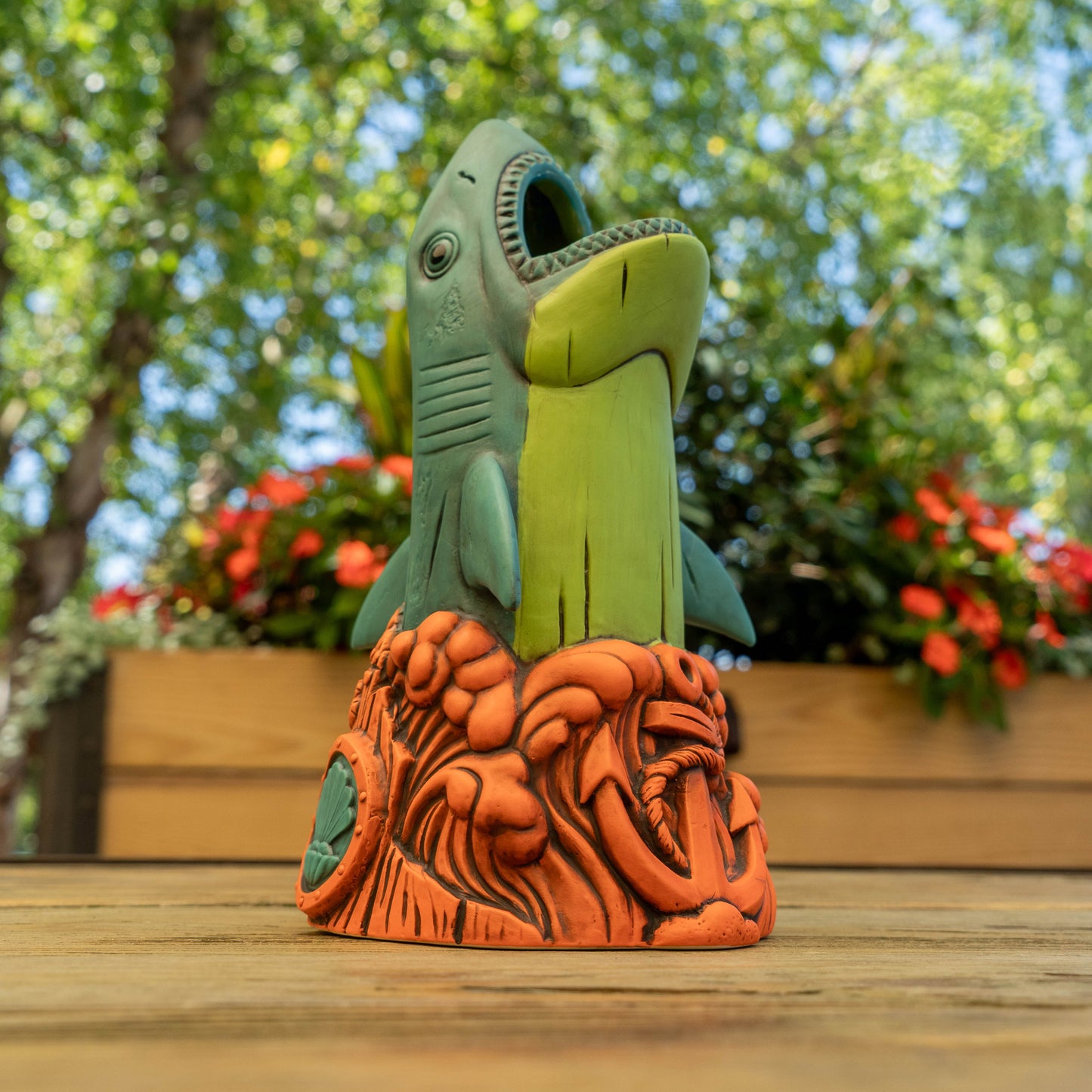 A shark-shaped ceramic cup with an open mouth, standing on an orange textured base. The shark is light green and teal, set against a blurred outdoor garden background with trees and flowers.