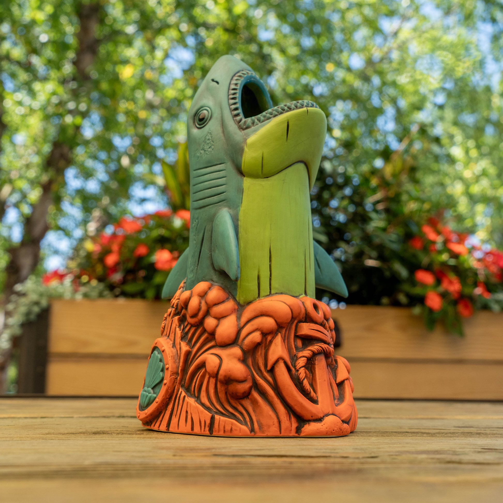 A shark-shaped ceramic cup with an open mouth, standing on an orange textured base. The shark is light green and teal, set against a blurred outdoor garden background with trees and flowers.