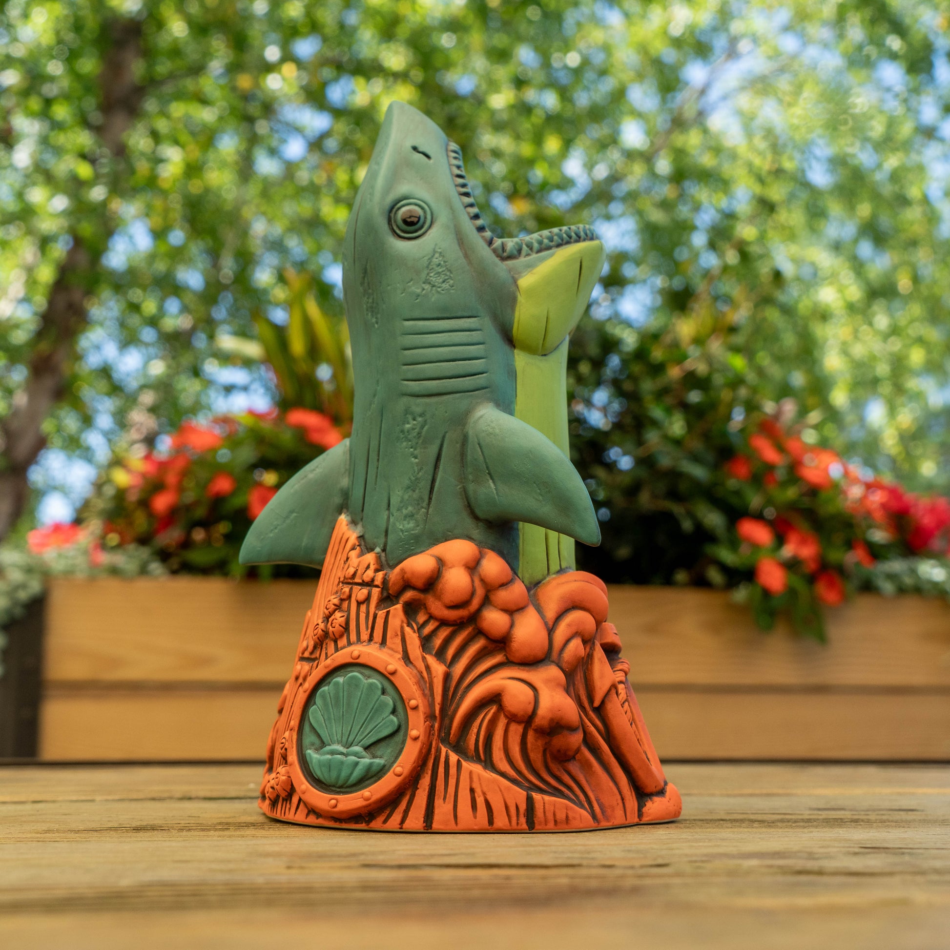 A shark cup with a green shark rising from an orange rock base, photographed outdoors on a wooden surface with blurred greenery and flowers in the background.