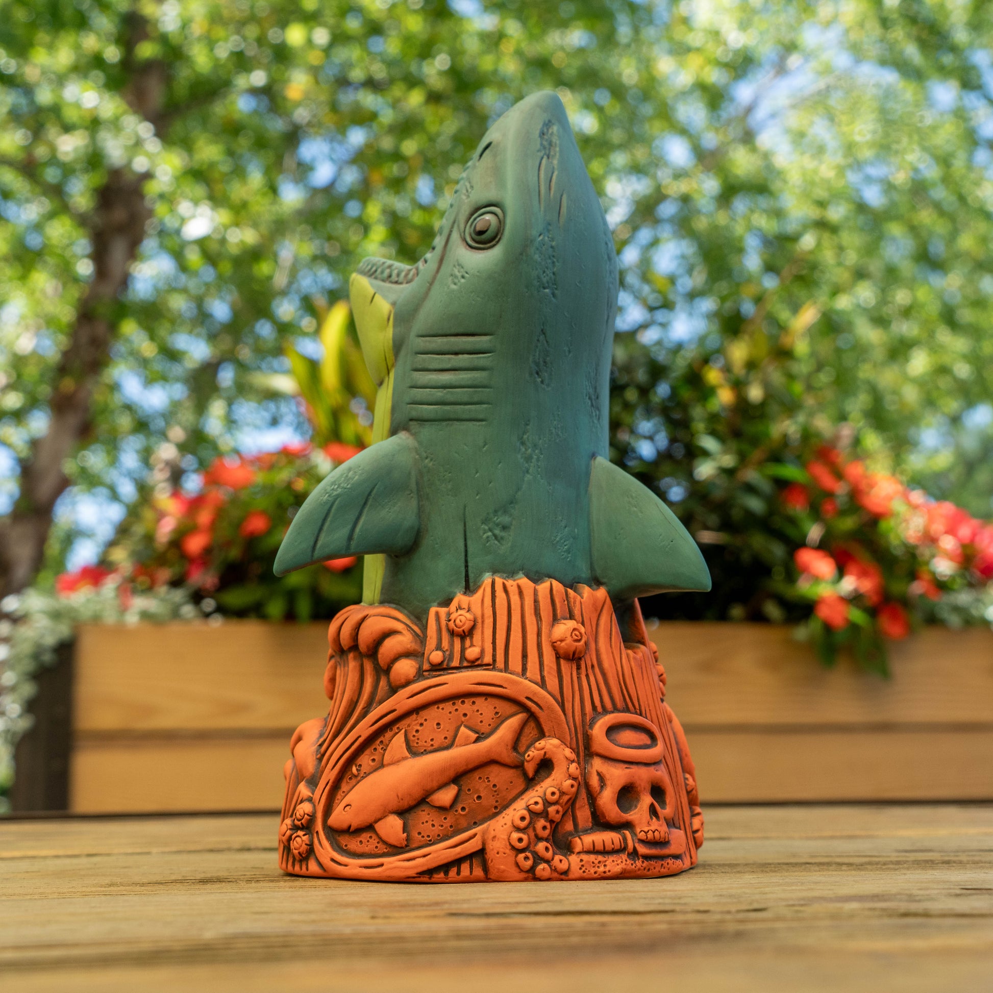 A ceramic shark-shaped cup in teal green rising from an orange terracotta base with carved sea-themed details, photographed outdoors on a wooden surface with blurred greenery and flowers in the background.