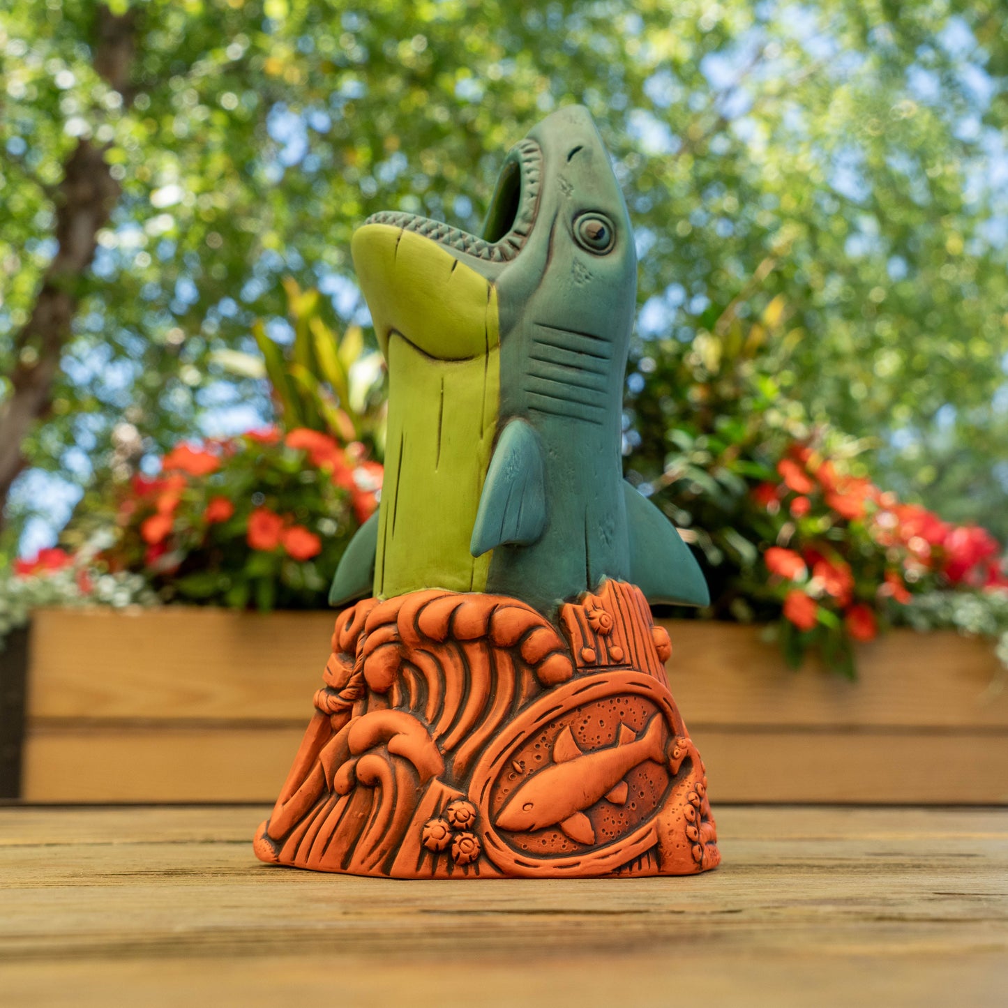 A colorful shark-shaped ceramic cup stands on an orange wave-patterned base outdoors on a wooden surface, with blurred greenery and red flowers in the background.
