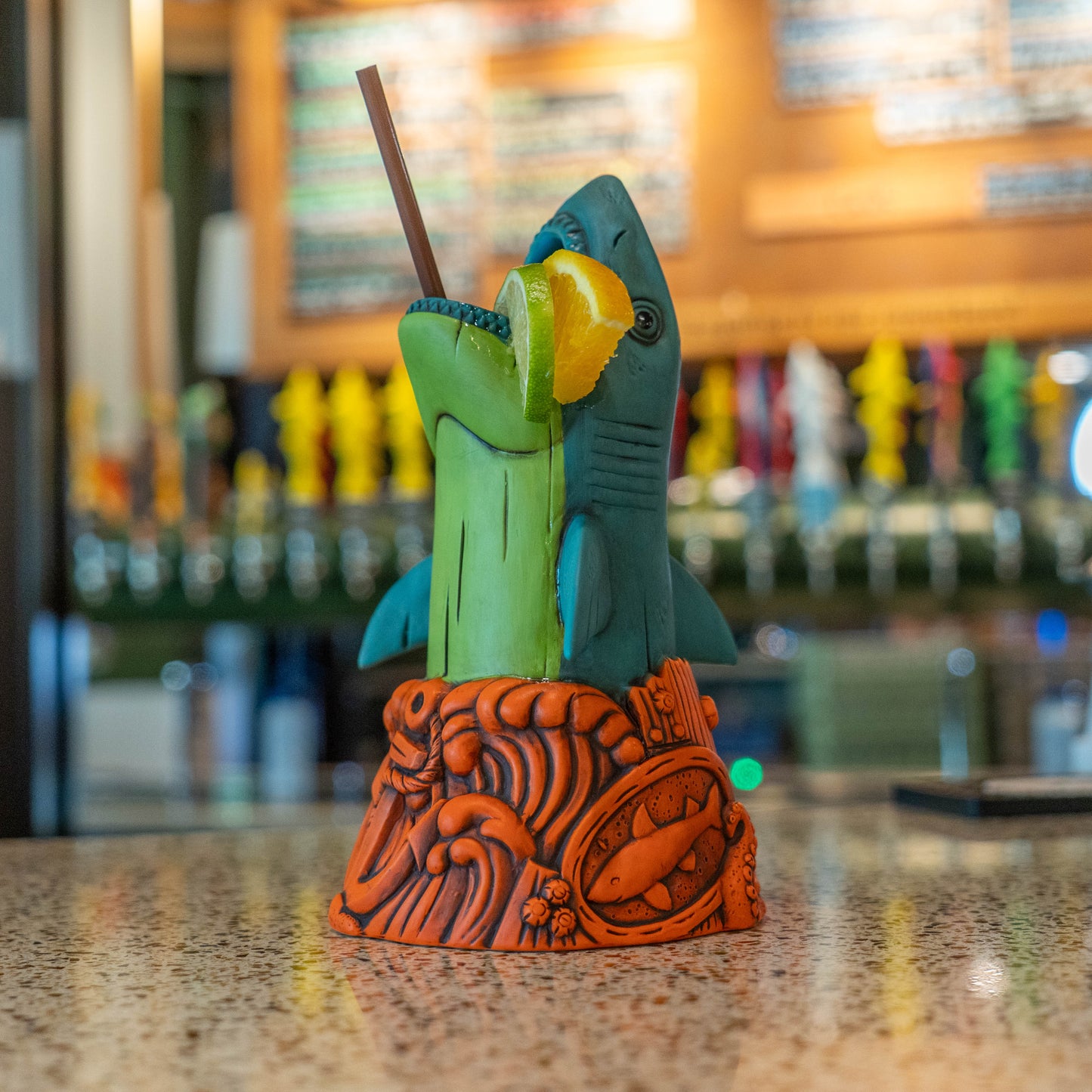 A colorful tropical shark cocktail holder on a speckled bar counter, with a green and blue shark, a yellow citrus slice, and an orange carved base, set against a blurred bar background.