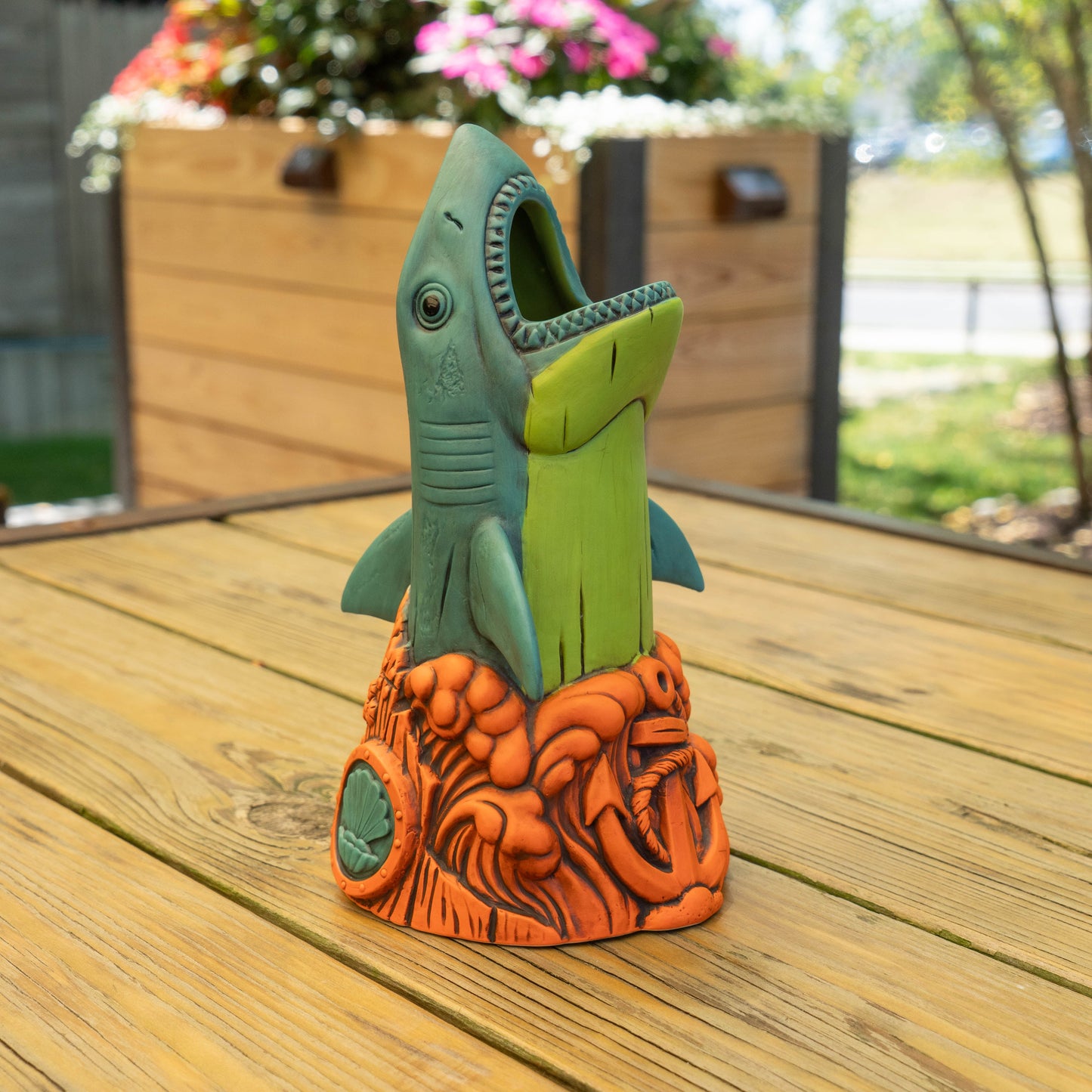 A decorative shark-shaped ceramic glass  sitting on a wooden outdoor table, with a green and teal shark design emerging from an orange textured base.