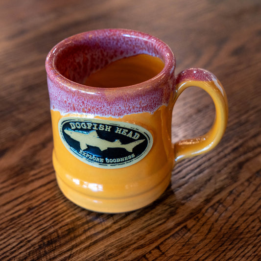 A Tangerine Ramsey Core Ceramic Mug by Deneen Pottery with a maroon rim sits on a wooden surface, featuring the Dogfish Head logo and "Explore Goodness" on its side.