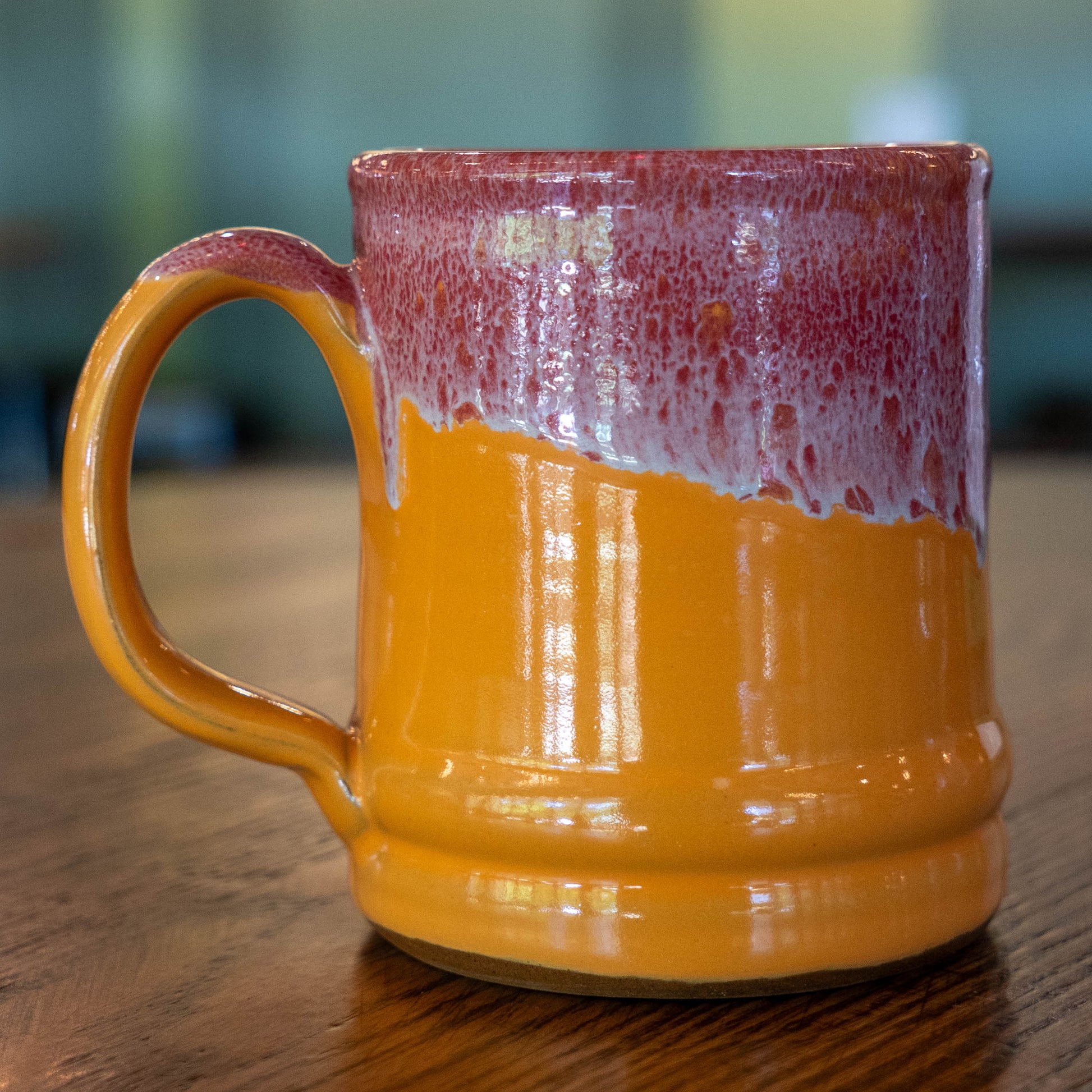 The Tangerine Ramsey Core Ceramic Mug by Deneen Pottery, featuring a glossy orange base and a red-and-white textured upper section, rests on a wooden table with a softly blurred background.