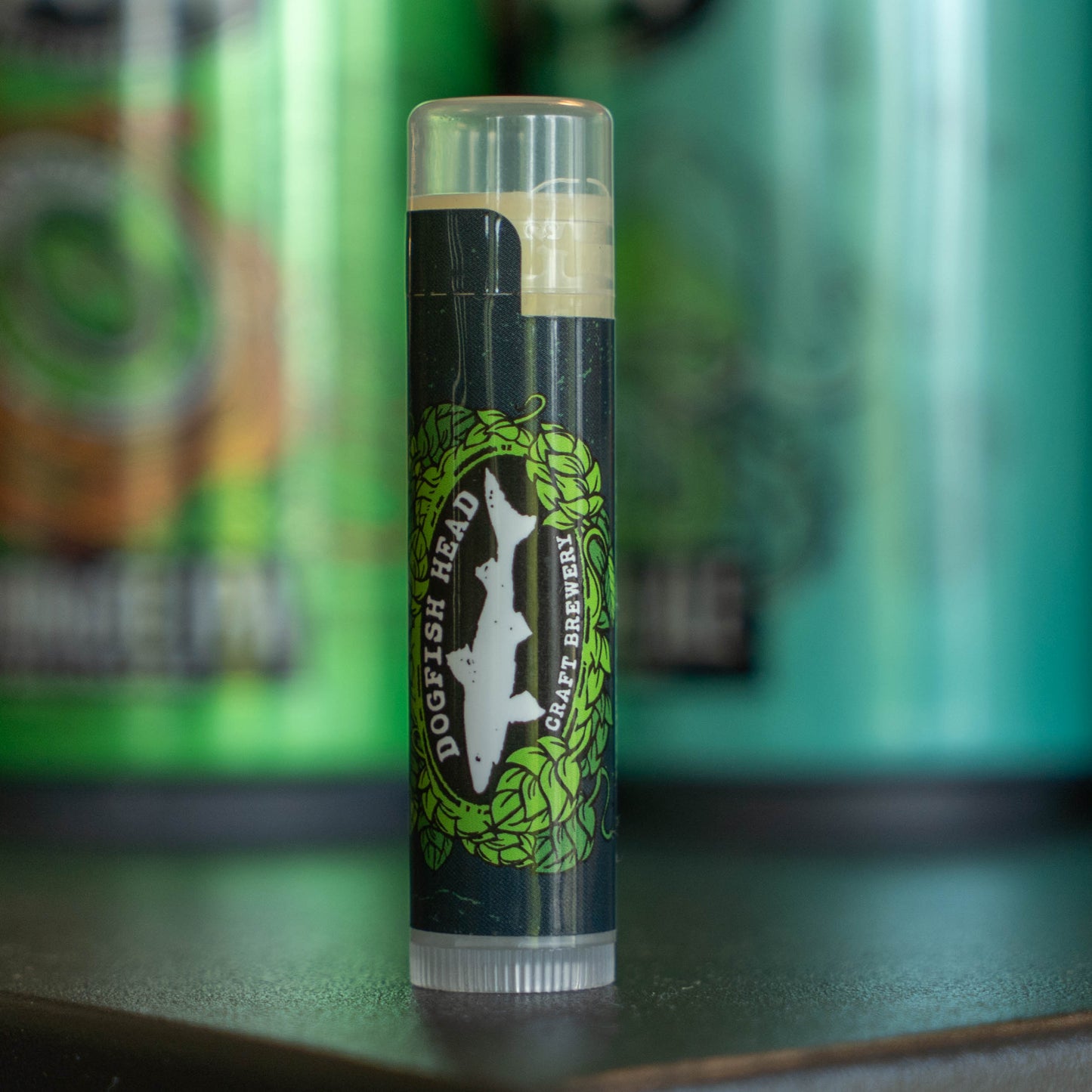 A tube of Ecolips Vanilla Lip Balm stands upright on a dark surface, with blurred green and black cans in the background.