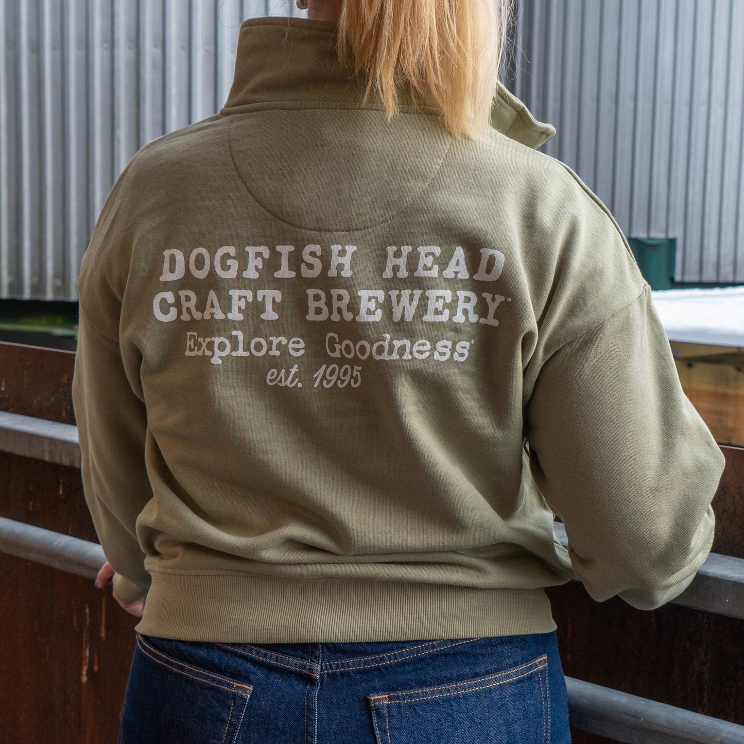 Back view of a woman wearing a Mineral quarter zip pullover sweatshirt with "Dogfish Head Craft Brewery, Explore Goodness, est. 1995" designed in white text.