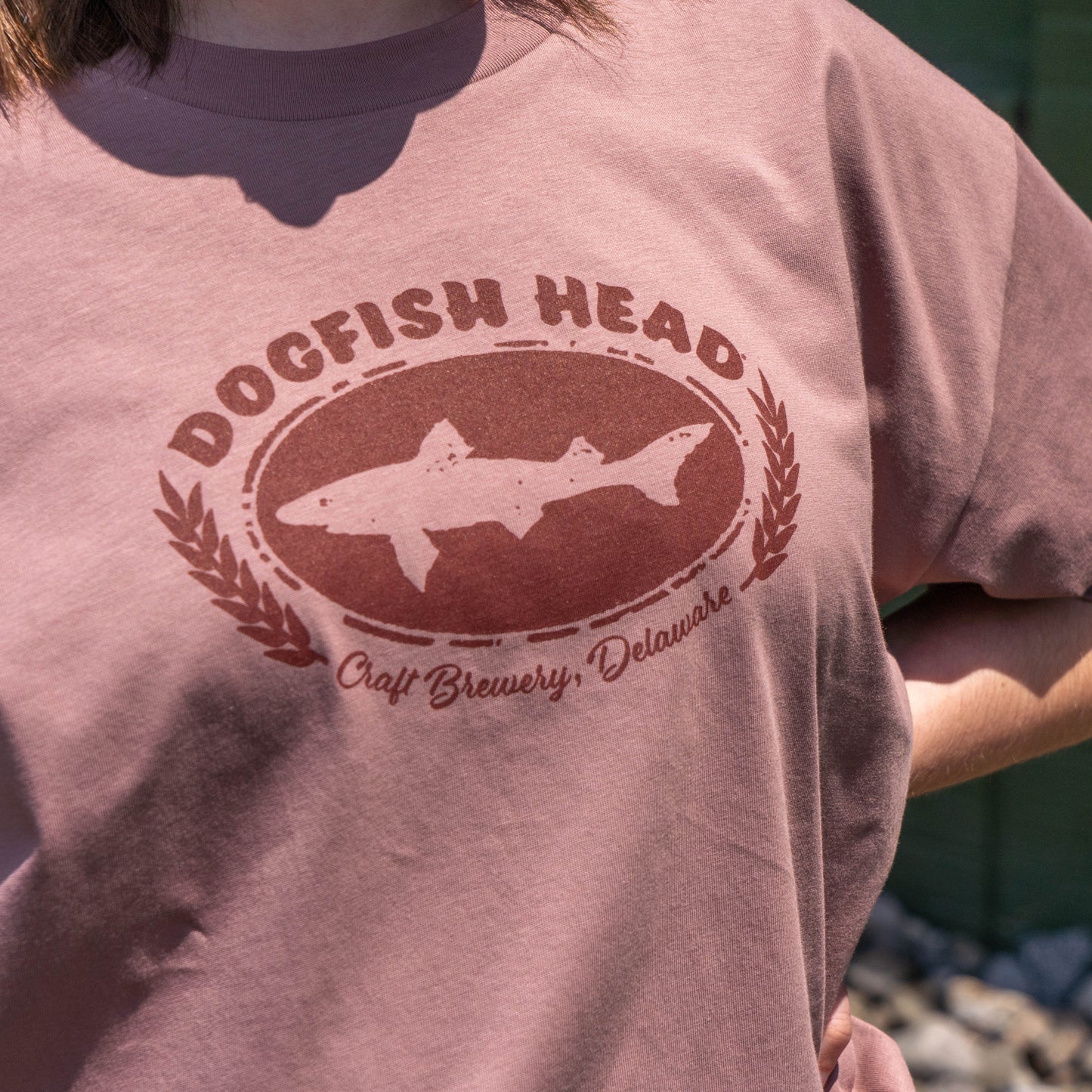 A person wears the W Pink Core Logo Tee Shirt by GO WEST T-SHIRT COMPANY, a women’s relaxed-fit mauve vintage-style tee with a Dogfish Head Craft Brewery, Delaware logo, featuring a stylized fish and laurel branches, outdoors in sunlight.