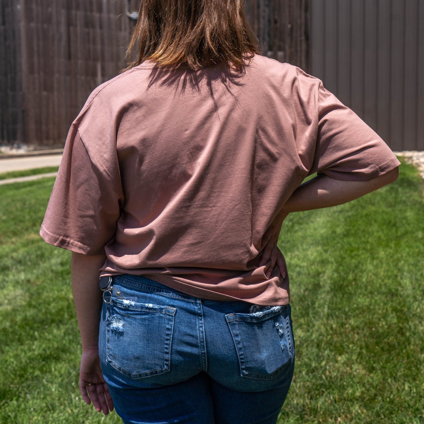 A person stands outdoors on grass with their back to the camera, wearing the W Pink Core Logo Tee Shirt by GO WEST T-SHIRT COMPANY and blue distressed jeans. One hand is on their hip, with buildings visible in the background.
