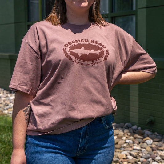 A woman in a relaxed-fit GO WEST T-SHIRT COMPANY W Pink Core Logo Tee Shirt and blue jeans stands outside near a green building and rocks on a sunny day.