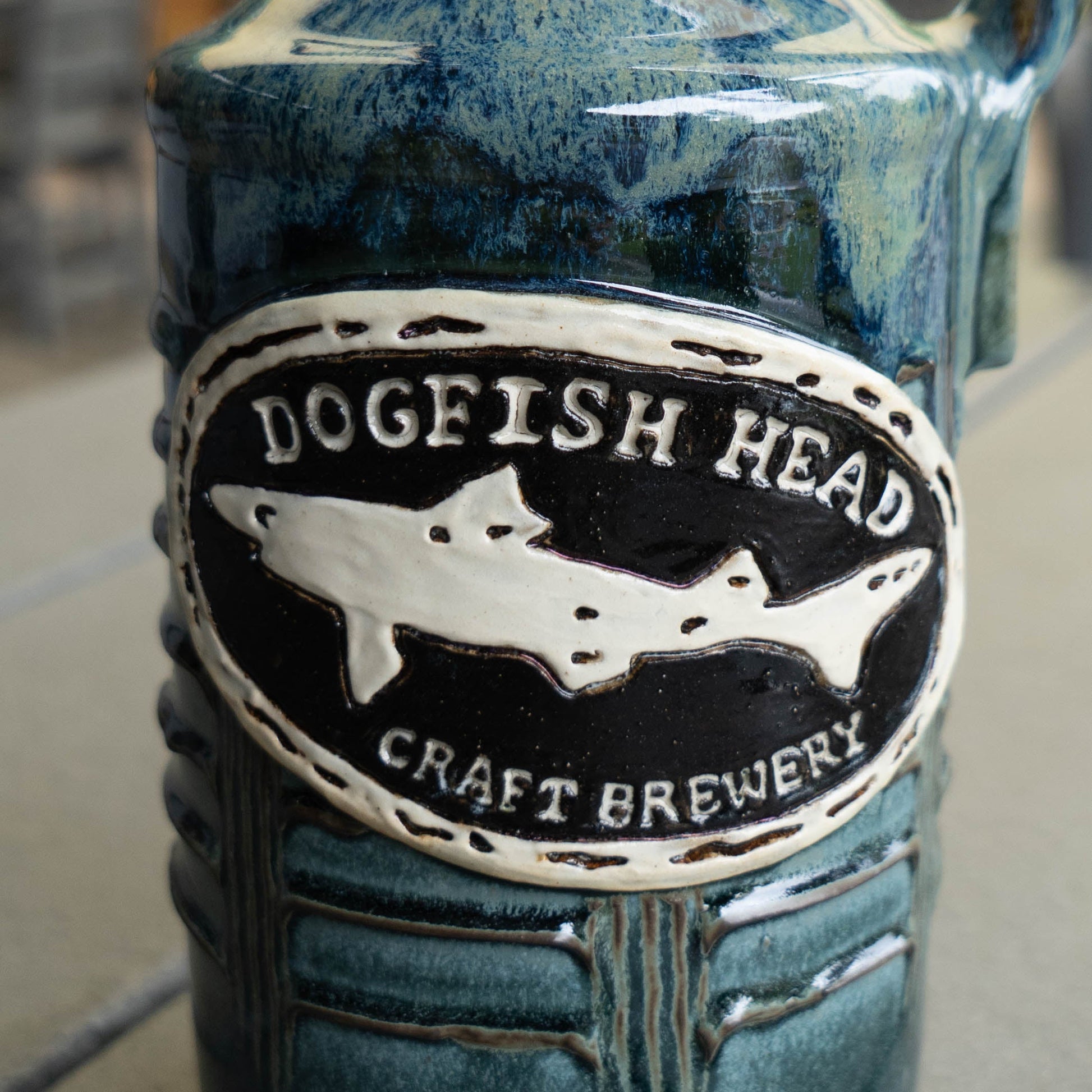 Close-up of the White Blue Green Ceramic Growler by Romanick Pottery, a hand-crafted jug glazed in blue and green shades. Features an oval logo embossed with Dogfish Head Craft Brewery and a shark silhouette.