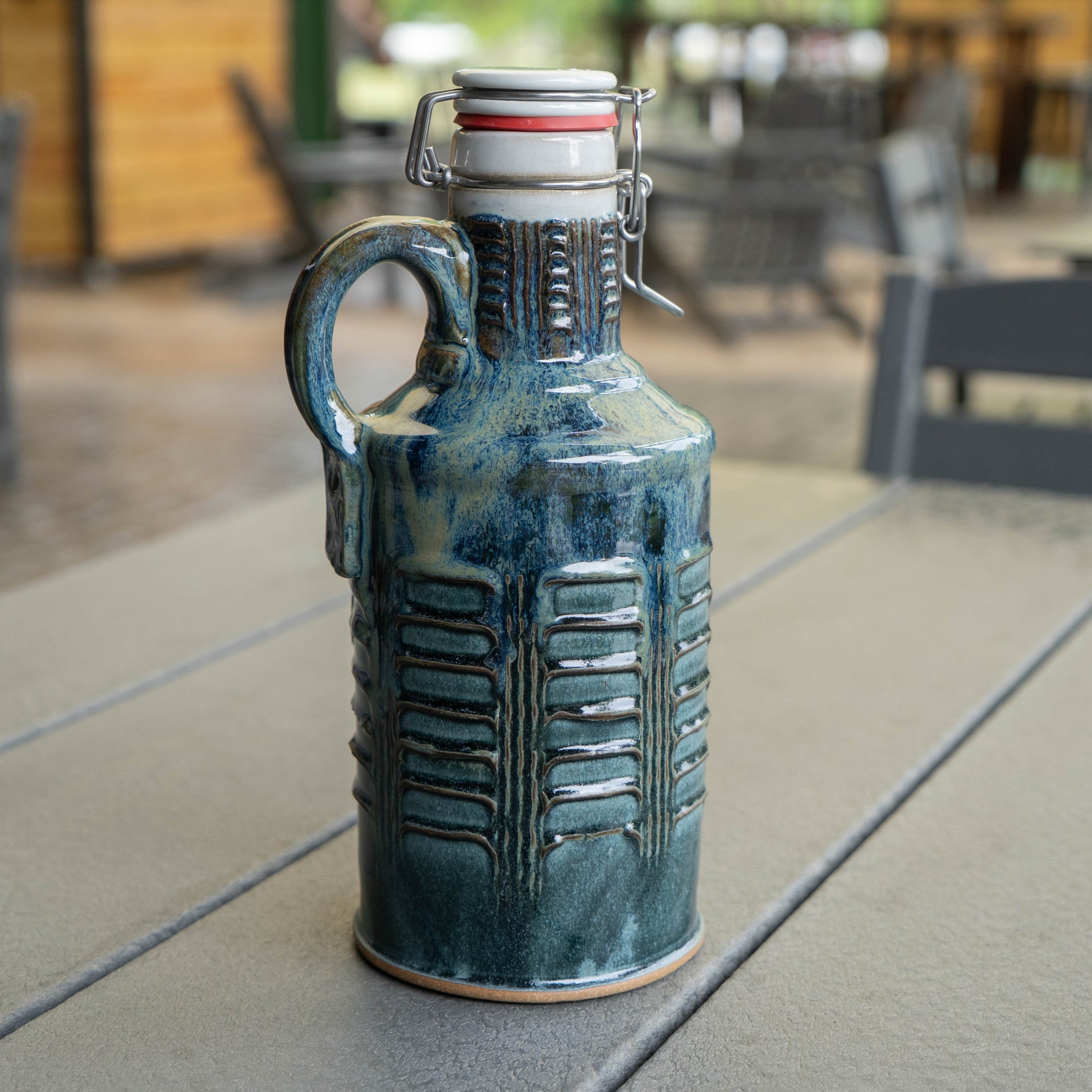The White Blue Green Ceramic Growler by Romanick Pottery features a glossy blue-green glaze, textured vertical patterns, and a flip-top lid with handle. It sits on an outdoor table, with blurred patio chairs and wooden walls in the background.