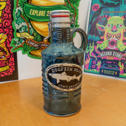A handmade White Blue Green Ceramic Growler by Romanick Pottery with a handle rests on a wooden surface, while colorful, vibrant posters decorate the background, enhancing the charm of this artisanal brew scene.