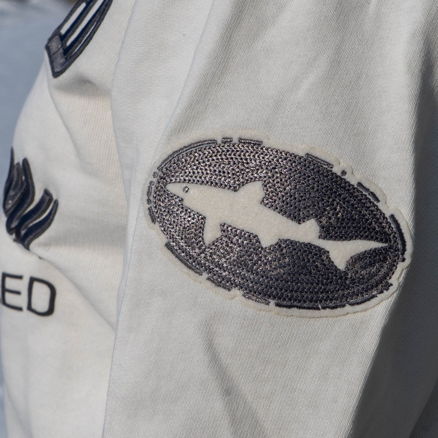 Close up of black embroidered dogfish head logo patch on the bicep of white off centered crew neck 