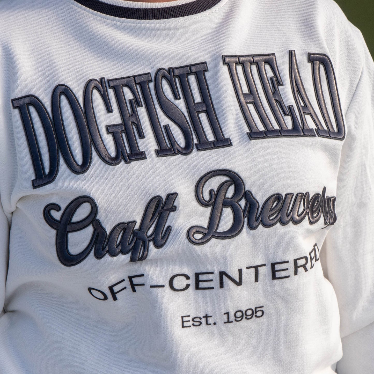 Close up of logo on white off centered crew neck.  Crew reads "Dogfish Head, Craft Brewery, OFF-CENTERED, Est. 1995"