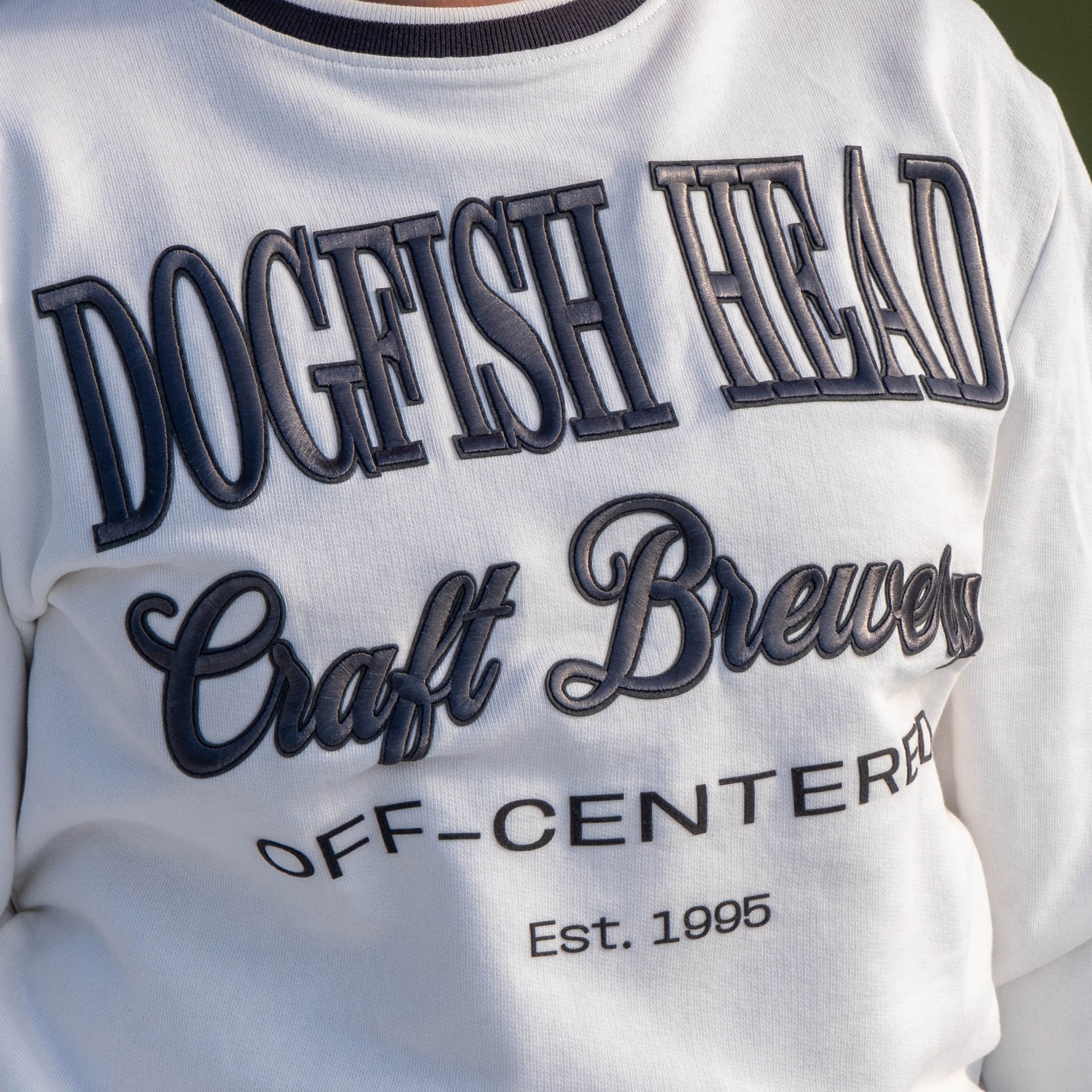 Close up of logo on white off centered crew neck.  Crew reads "Dogfish Head, Craft Brewery, OFF-CENTERED, Est. 1995"