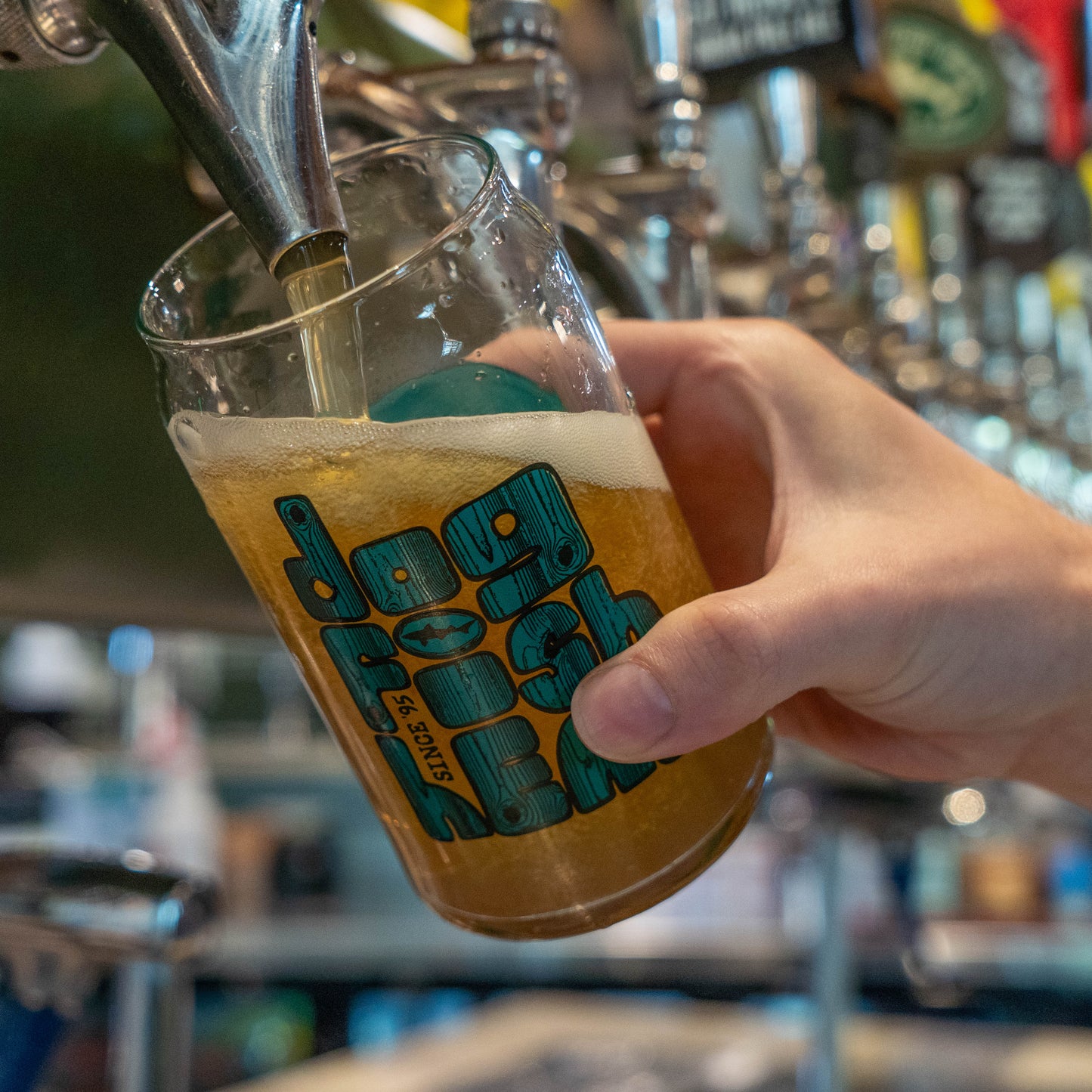 Hand pouring beer into can glass with a dogfish head logo styled in teal and wood grain