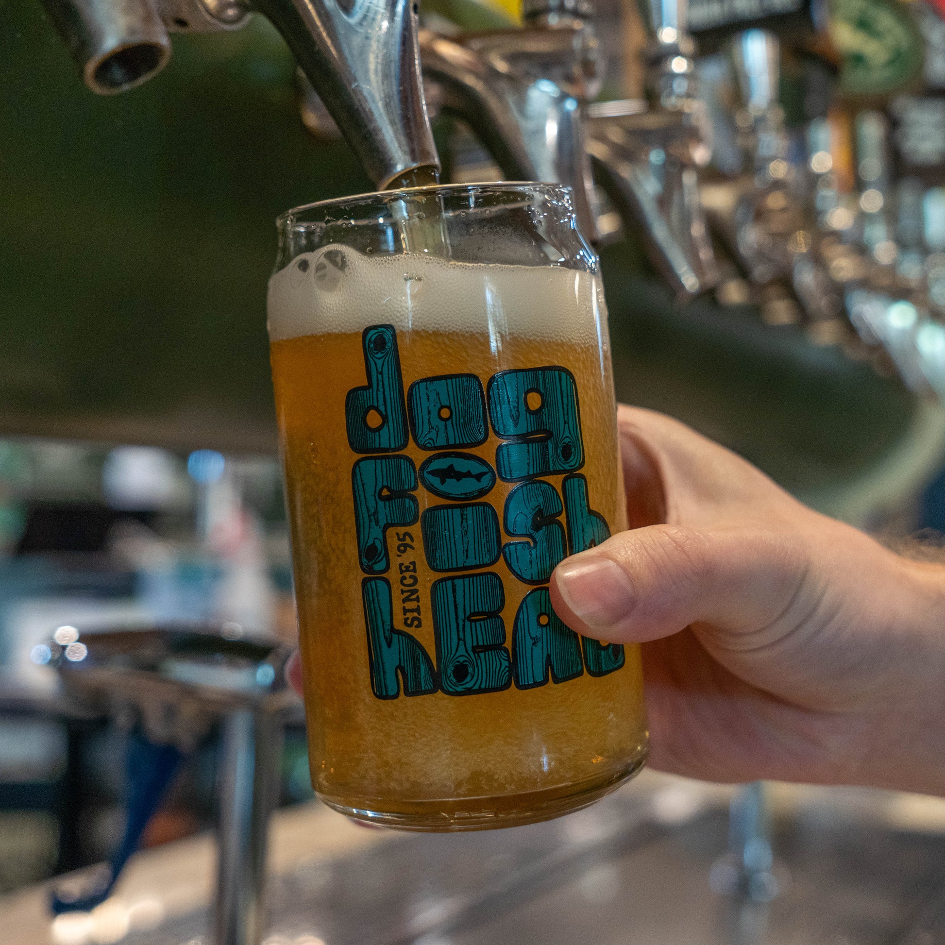 Hand pouring beer into can glass with a dogfish head logo styled in teal and wood grain