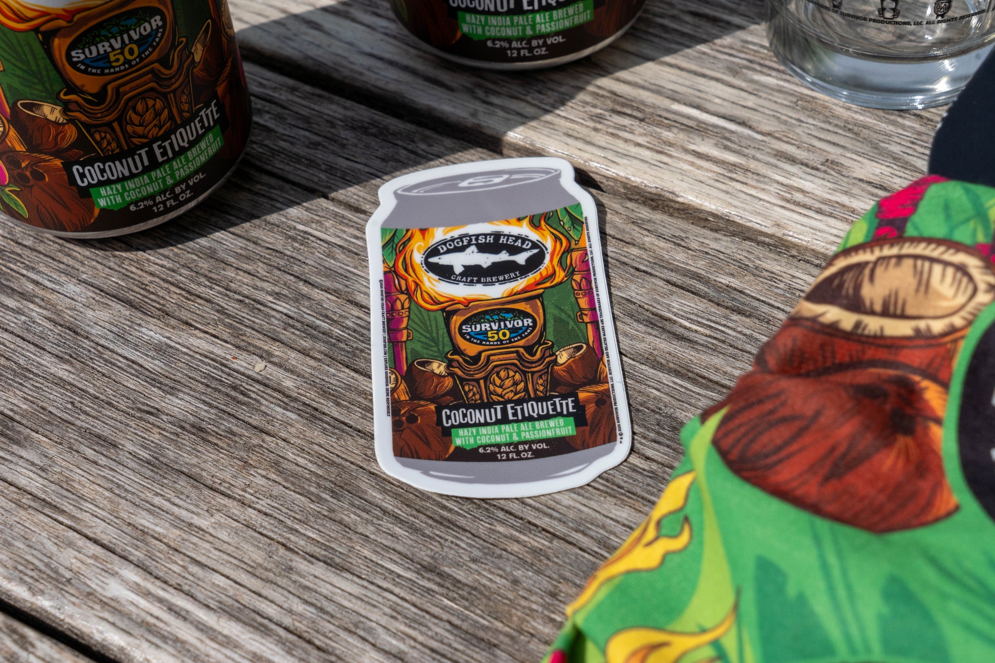 A sticker from the Dogfish Head x Survivor 50 Bundle (MERCH ONLY) by Grandstand Glassware & Apparel, shaped like a soda can with Coconut Etiquette Survivor 50 art, rests on a wooden table near a beer can and colorful fabric.