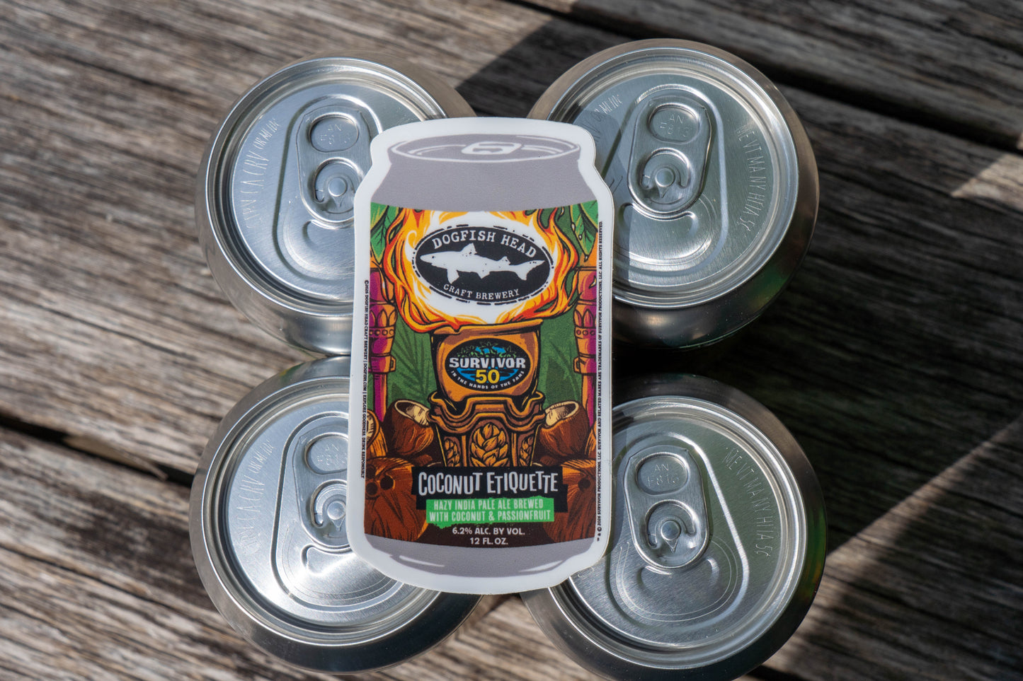 Four silver cans rest on wood, adorned with a vibrant Dogfish Head x Survivor 50 Bundle label from Grandstand Glassware & Apparel. The tropical, tiki-inspired design nods to exclusive Survivor 50th season merch, partially covering the tops.