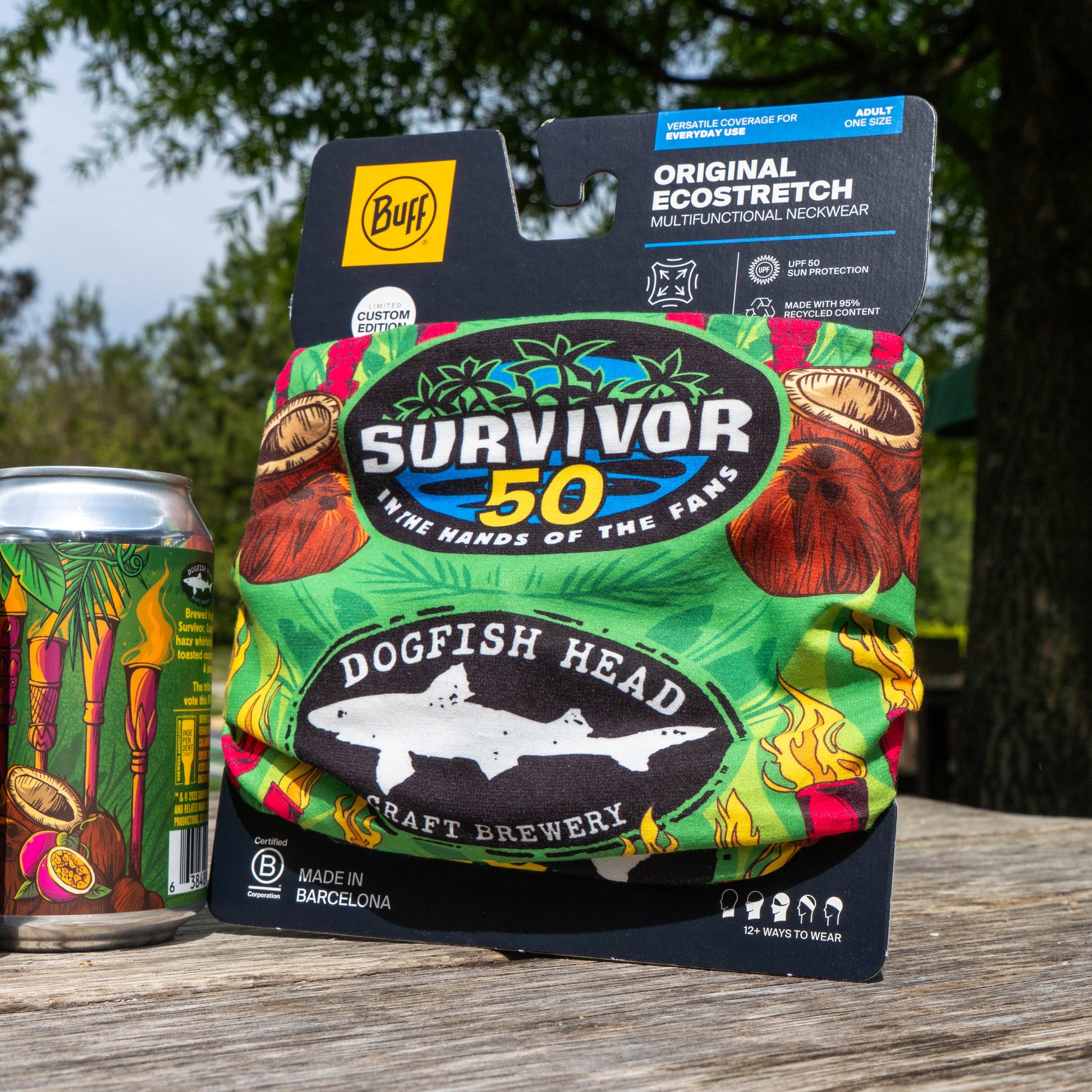 The Dogfish Head x Survivor 50 Bundle (MERCH ONLY) by Grandstand Glassware & Apparel, a limited edition collectible, sits on a wooden table outdoors beside a can of beer, with trees and grass softly blurred in the background.