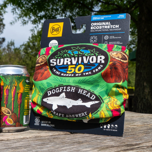 The Dogfish Head x Survivor 50 Bundle (MERCH ONLY) by Grandstand Glassware & Apparel, a limited edition collectible, sits on a wooden table outdoors beside a can of beer, with trees and grass softly blurred in the background.