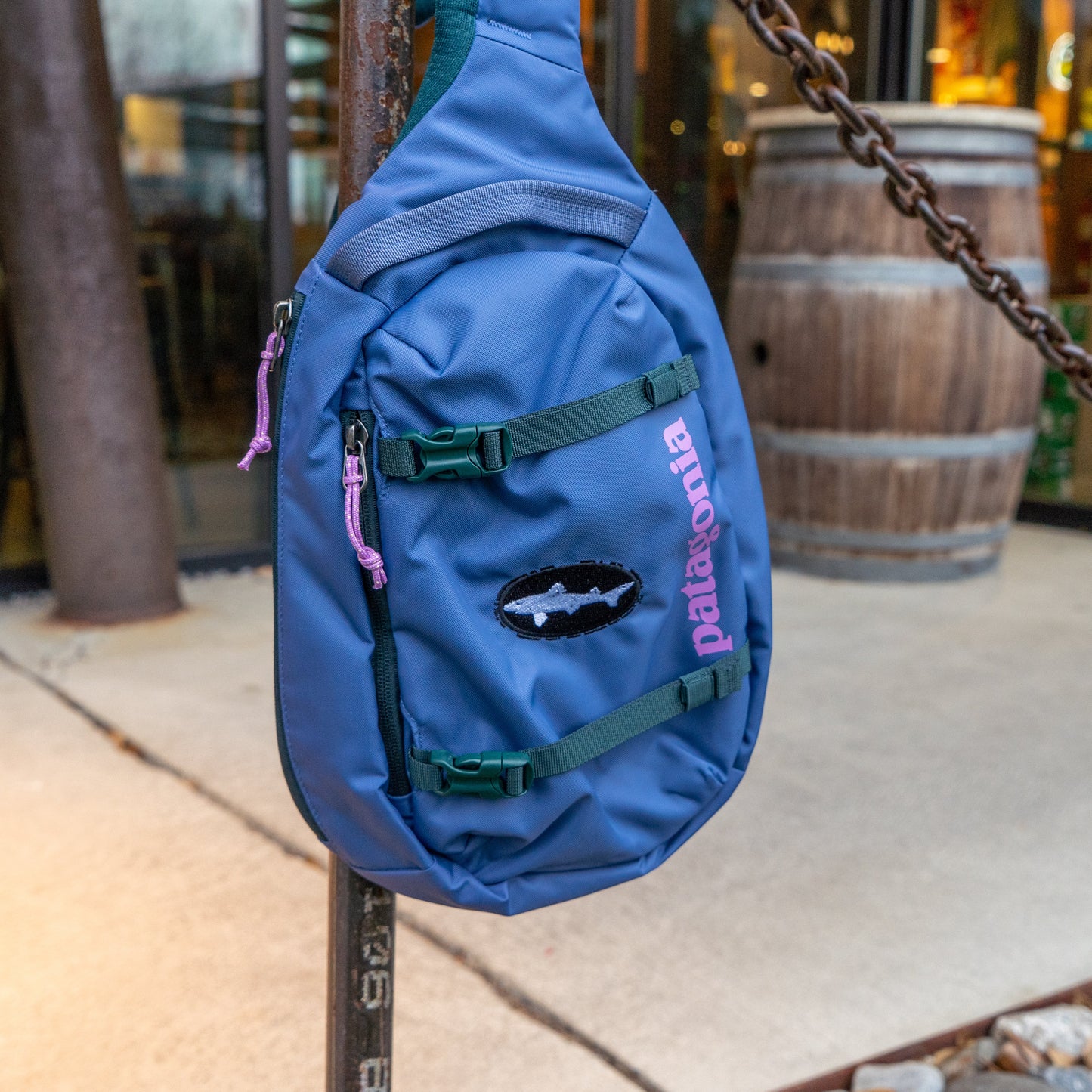 The Patagonia Blue Atom Sling 8L, made from recycled polyester, hangs from a rusty metal post outdoors with a wooden barrel and glass doors in the background.