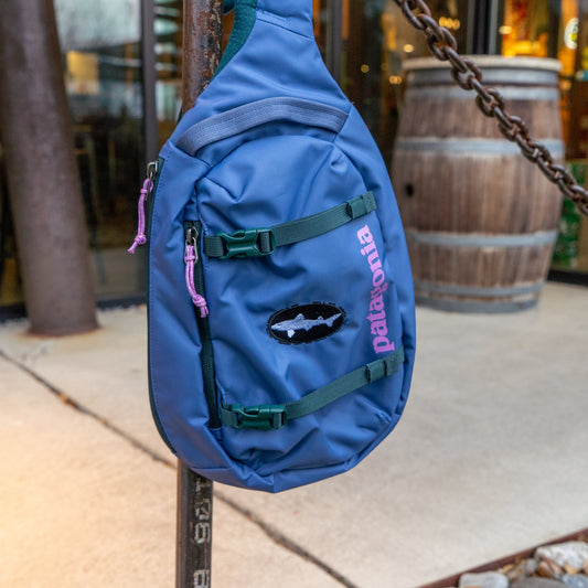 The Patagonia Blue Atom Sling 8L, made from recycled polyester, hangs from a rusty metal post outdoors with a wooden barrel and glass doors in the background.
