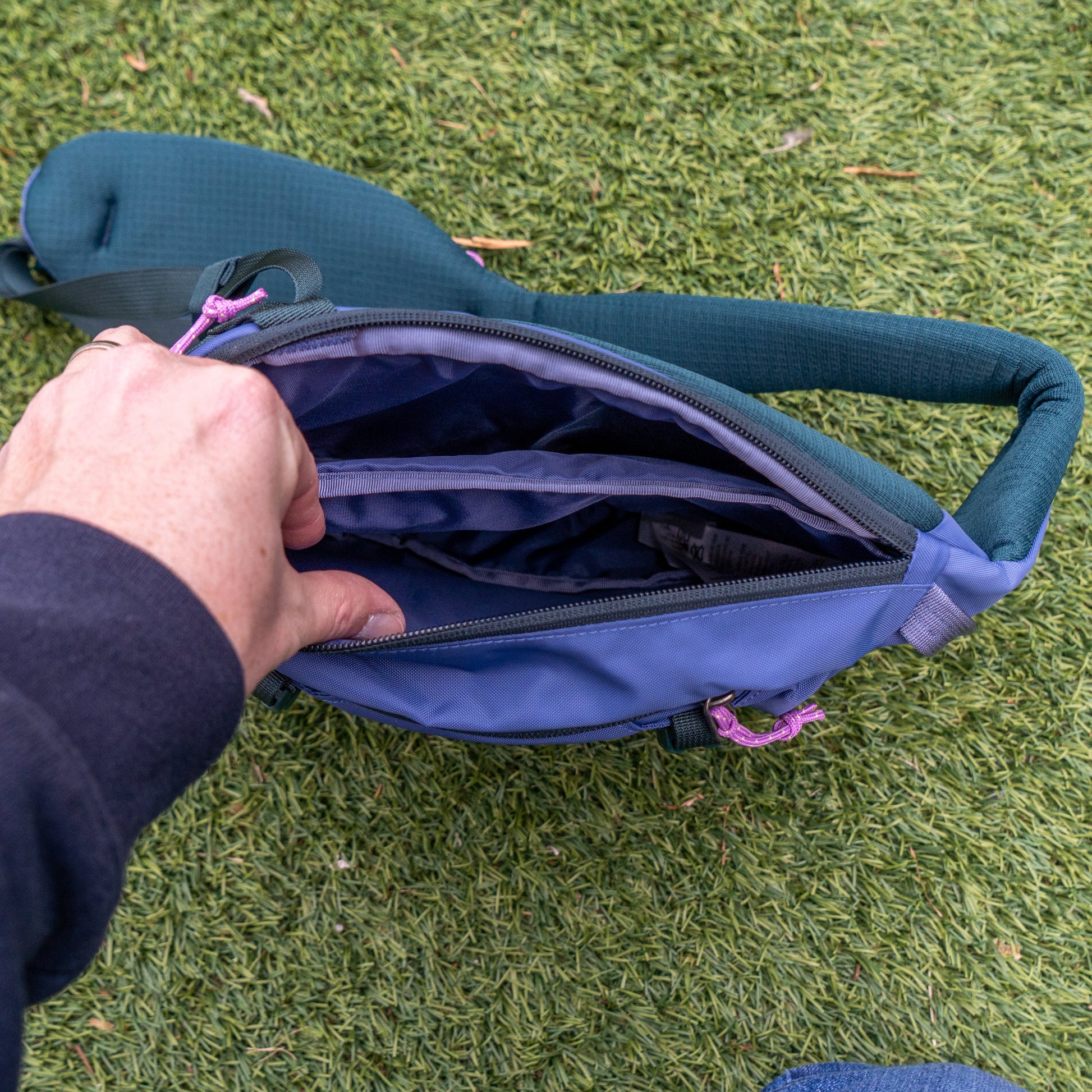 A person unzips a Patagonia Blue Atom Sling 8L, showing its multiple compartments. The blue bag rests on green artificial grass.