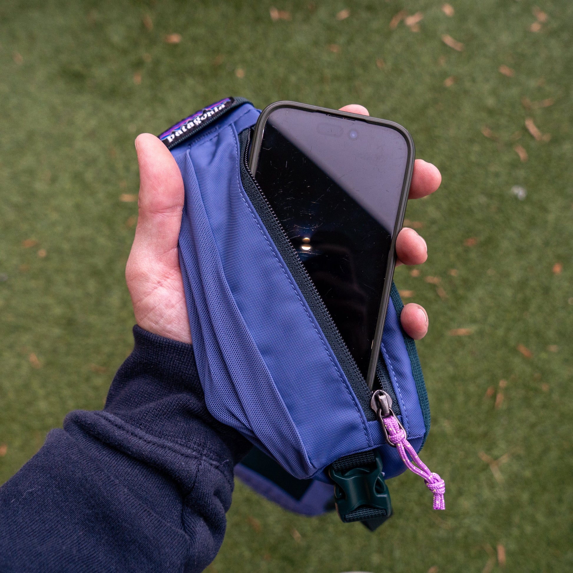 A hand holds a Patagonia Blue Atom Sling 8L, made with recycled polyester. A smartphone peeks from the pouch, which features a Patagonia tag and purple zipper pull. The grassy background is scattered with leaves.